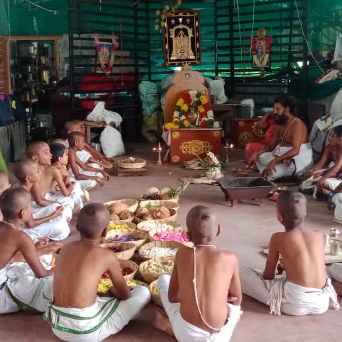 Students seated in a circle around baskets of coconuts, flowers and offerings while a priest conducts a homa before a decorated altar