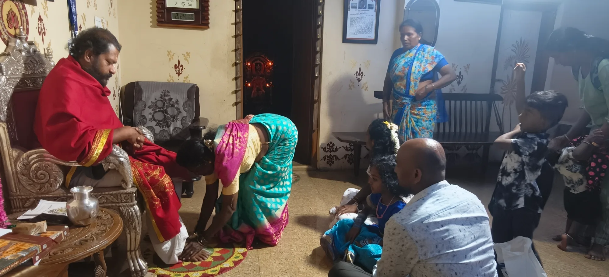 A woman bending to touch the feet of the vaidika seated in a red shawl at the end of a sitting, her husband and children waiting on the floor beside her with the chart and the vessels still laid out