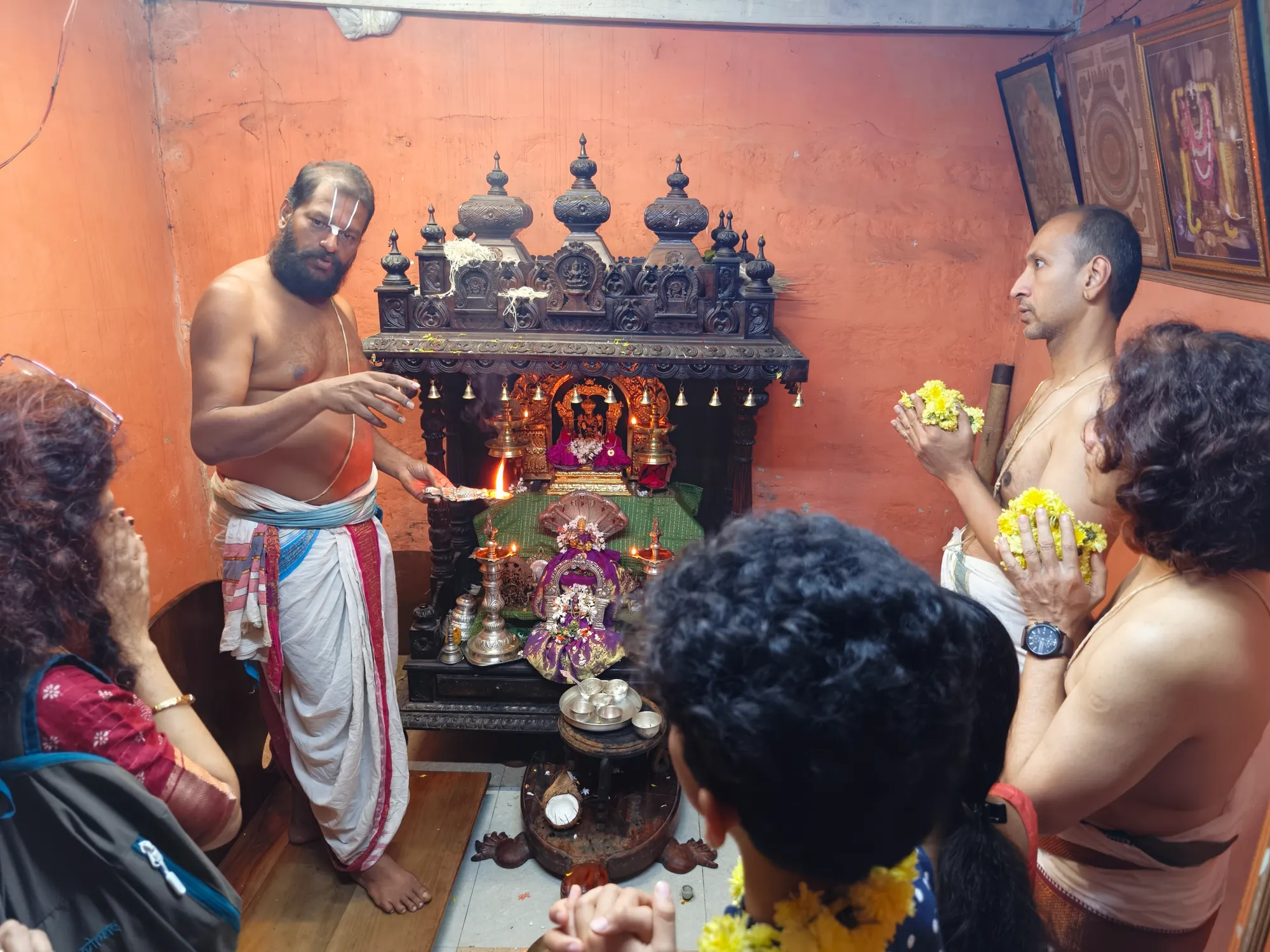 A vaidika standing at the shrine explaining a reading to the family who have come for it, the lamp lit before the deity and flowers held in their joined hands