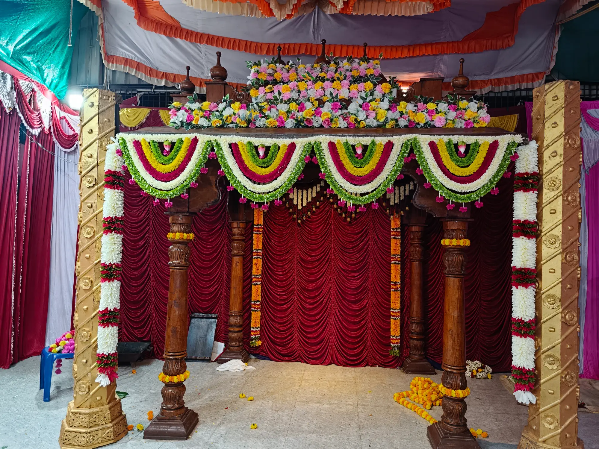 The mandapa dressed and still empty — carved wooden pillars, swags of white and green garlands, and a bank of flowers laid along the canopy