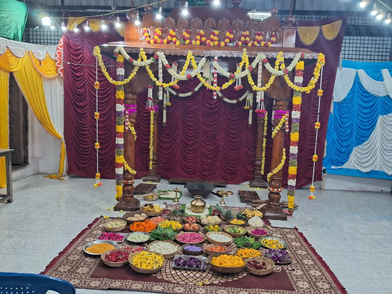 The carved wooden mandapa dressed with yellow garlands against a maroon curtain, the offerings ranged on a carpet at its foot