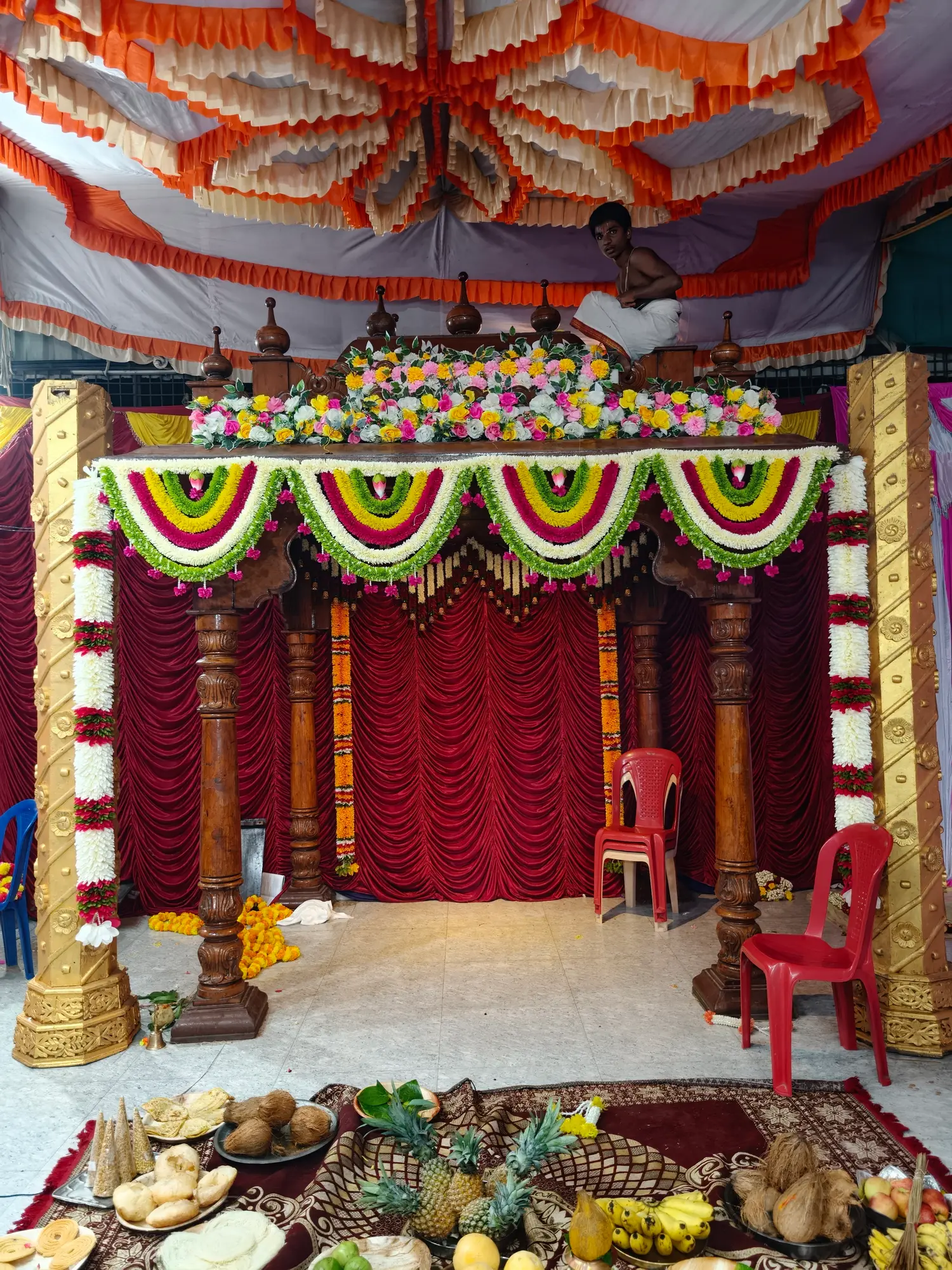 The mandapa from below, its roof banked in rows of white, pink and green garlands, the offerings set out on a carpet at the front