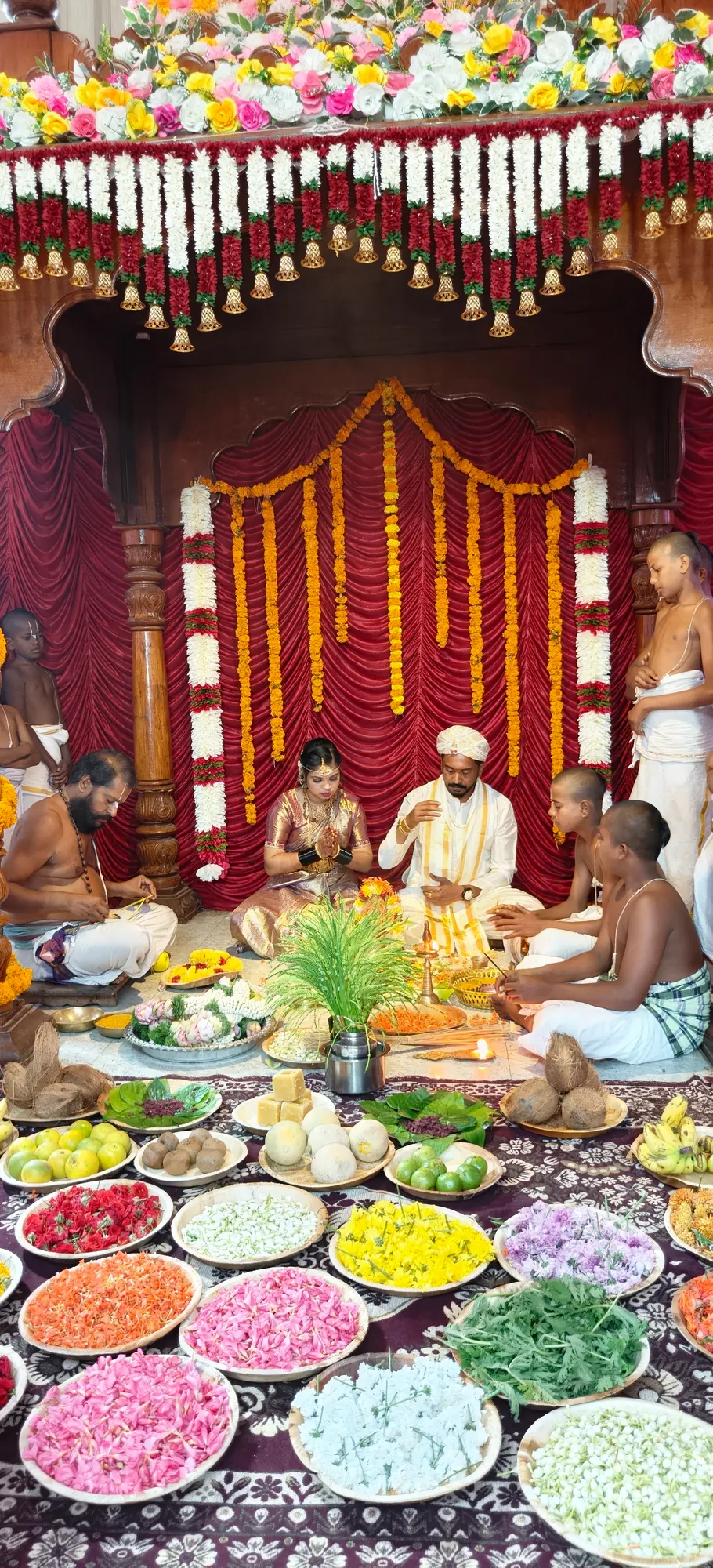 The kalyana mantapa seen end on — the couple seated under the carved wooden mandapa hung with marigold and jasmine, the priests around them, and the offering plates filling the whole foreground
