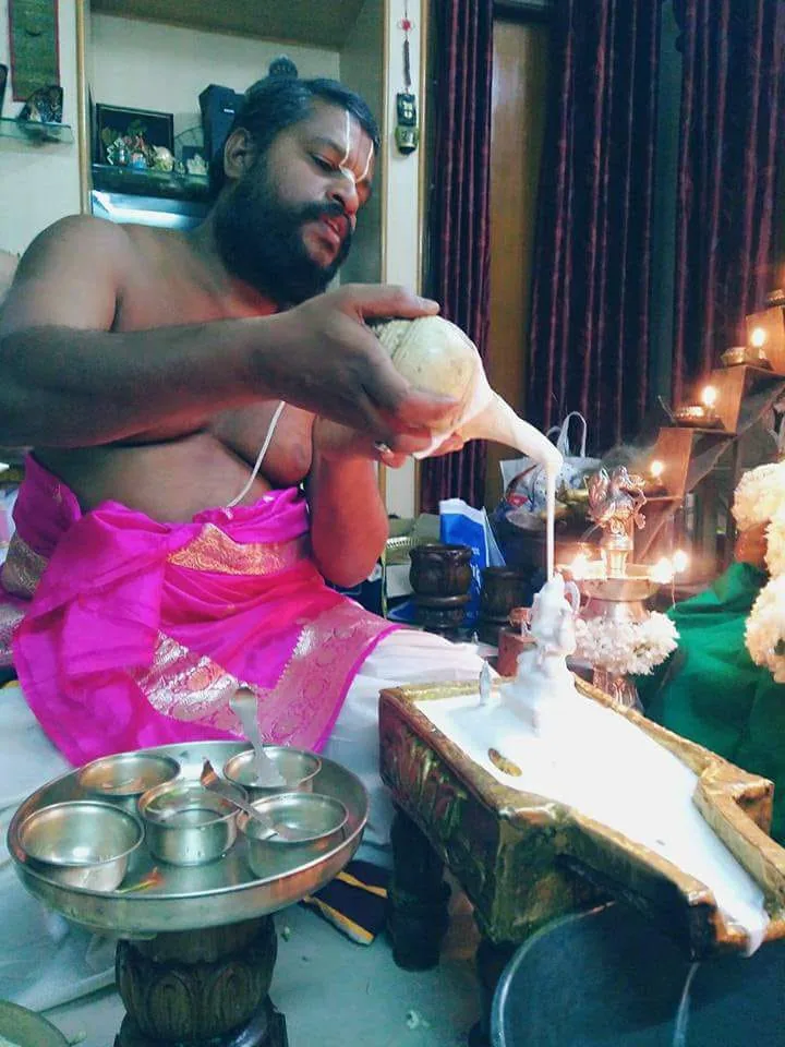 A vaidika in rose silk raising a conch over the murti to pour the abhisheka, the row of silver vessels laid out in front of him