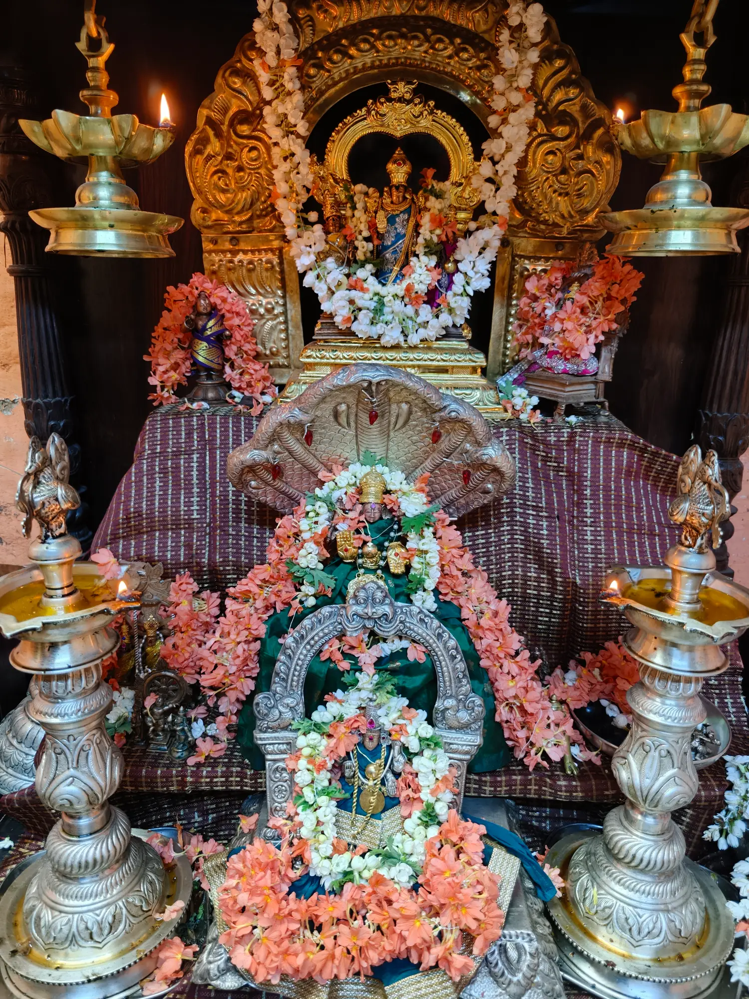 The full altar of the Gurupeeta pooja mandira — a gilded prabhavali above, a silver naga hood below it, and lit brass lamps standing at each corner, everything wound about with peach blossom and jasmine
