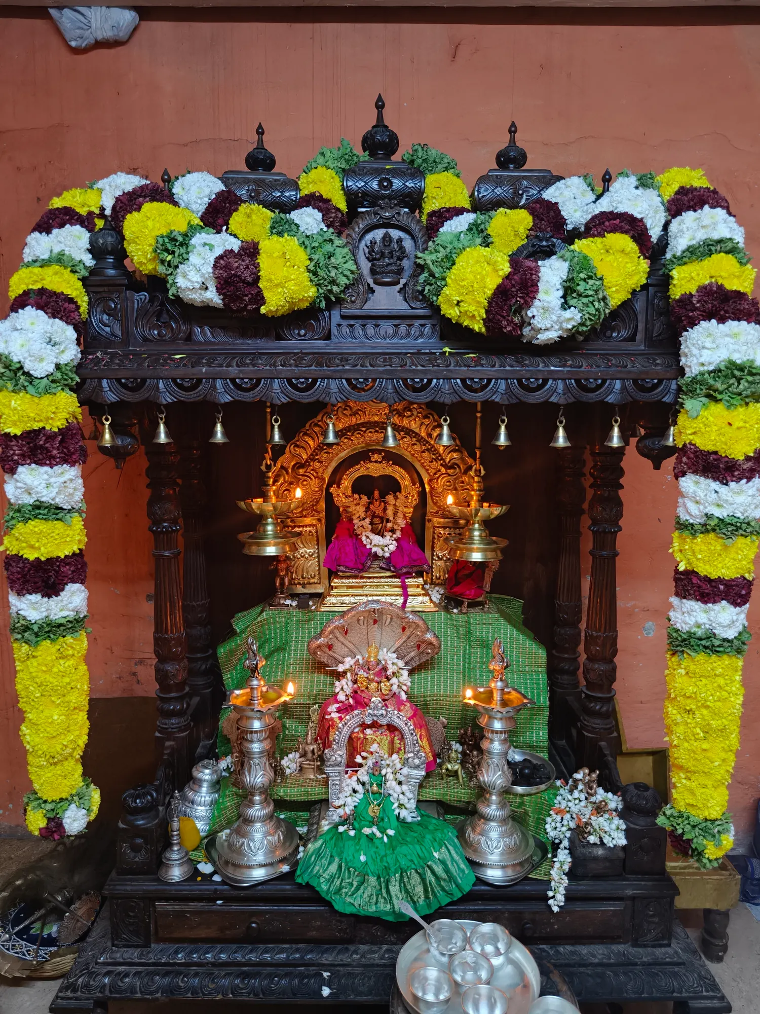 A carved dark wood shrine in the pooja mandira hung with rows of brass bells and thick garlands of marigold, white and purple flowers, lamps burning inside it