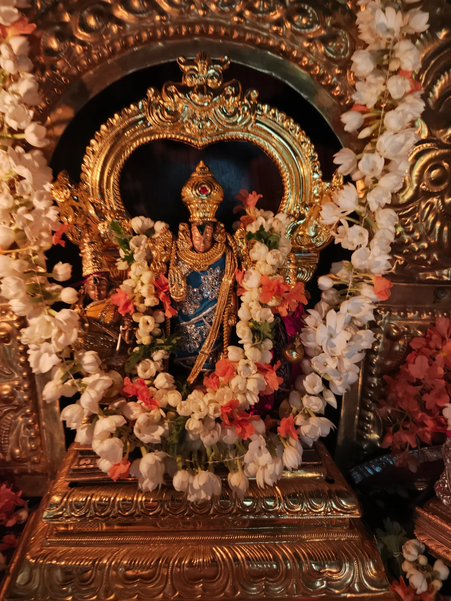 A small standing murti inside a golden prabhavali, framed by hanging strands of jasmine and coral-coloured blossom