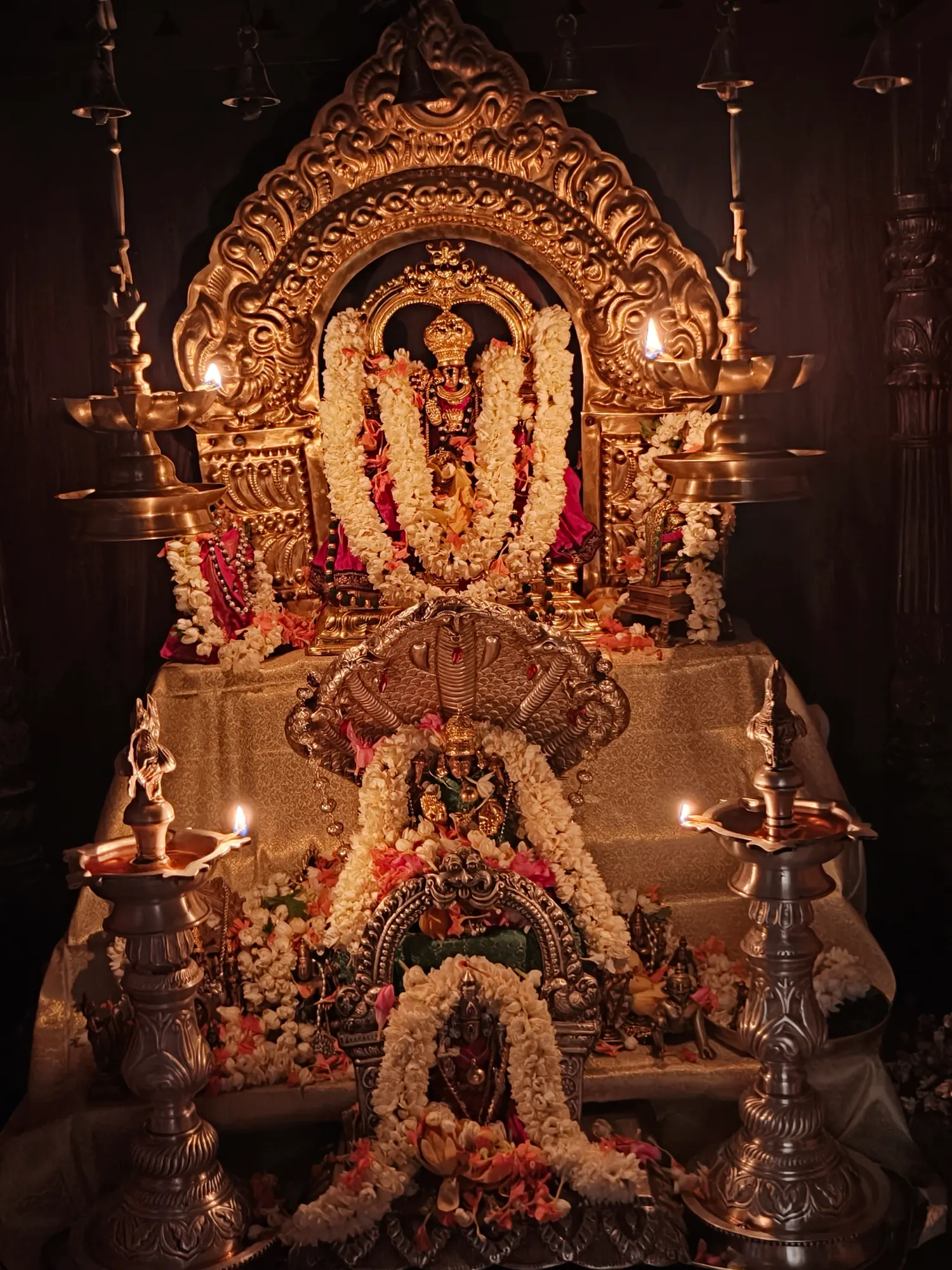 The altar of the pooja mandira in darkness, the gilded prabhavali and the deities beneath it lit only by the oil lamps burning on either side
