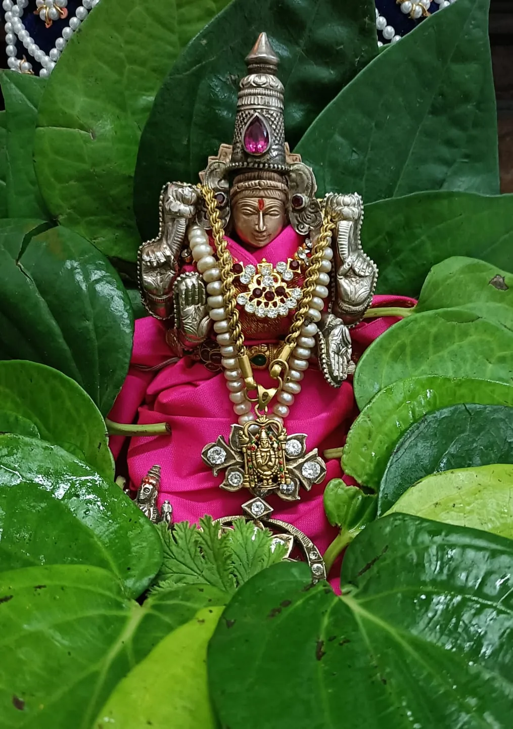 A silver murti with a pink gemstone crown set on fresh betel leaves and rose silk, gold and pearl garlands laid over it