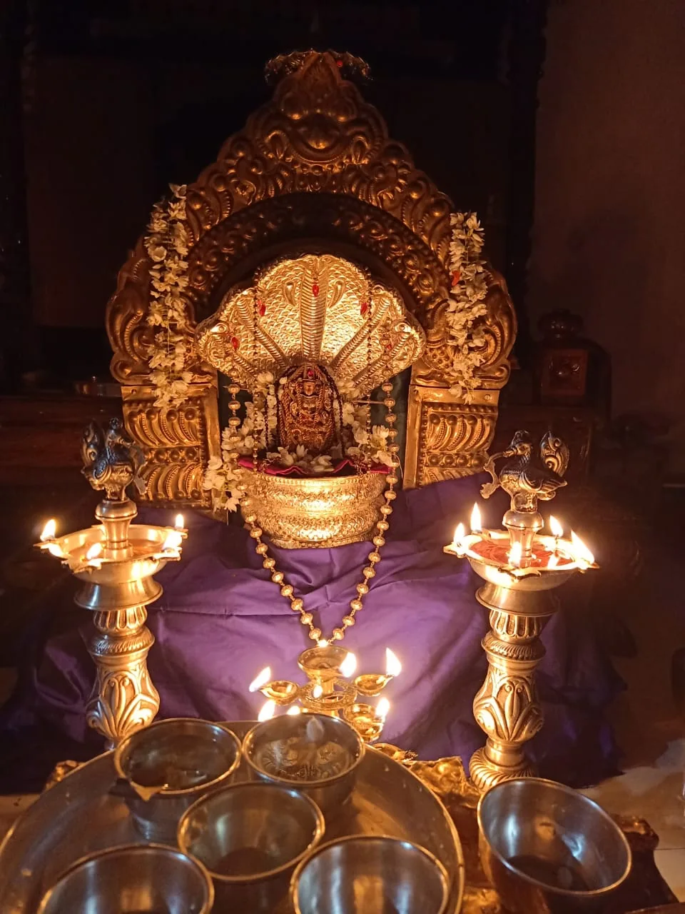 The gilded prabhavali of the mandira with a silver naga hood spread beneath it, tall brass lamps burning at either side and the offering vessels below