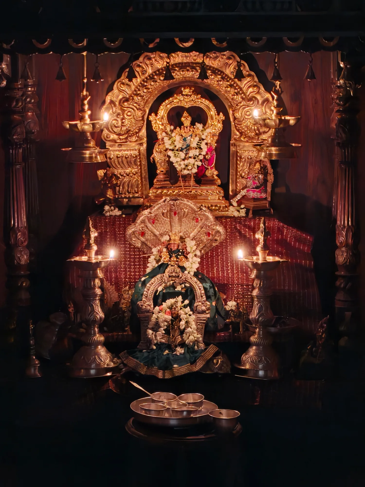 The carved wooden shrine of the mandira lit only by its own lamps, the gilded prabhavali above and a silver naga hood below