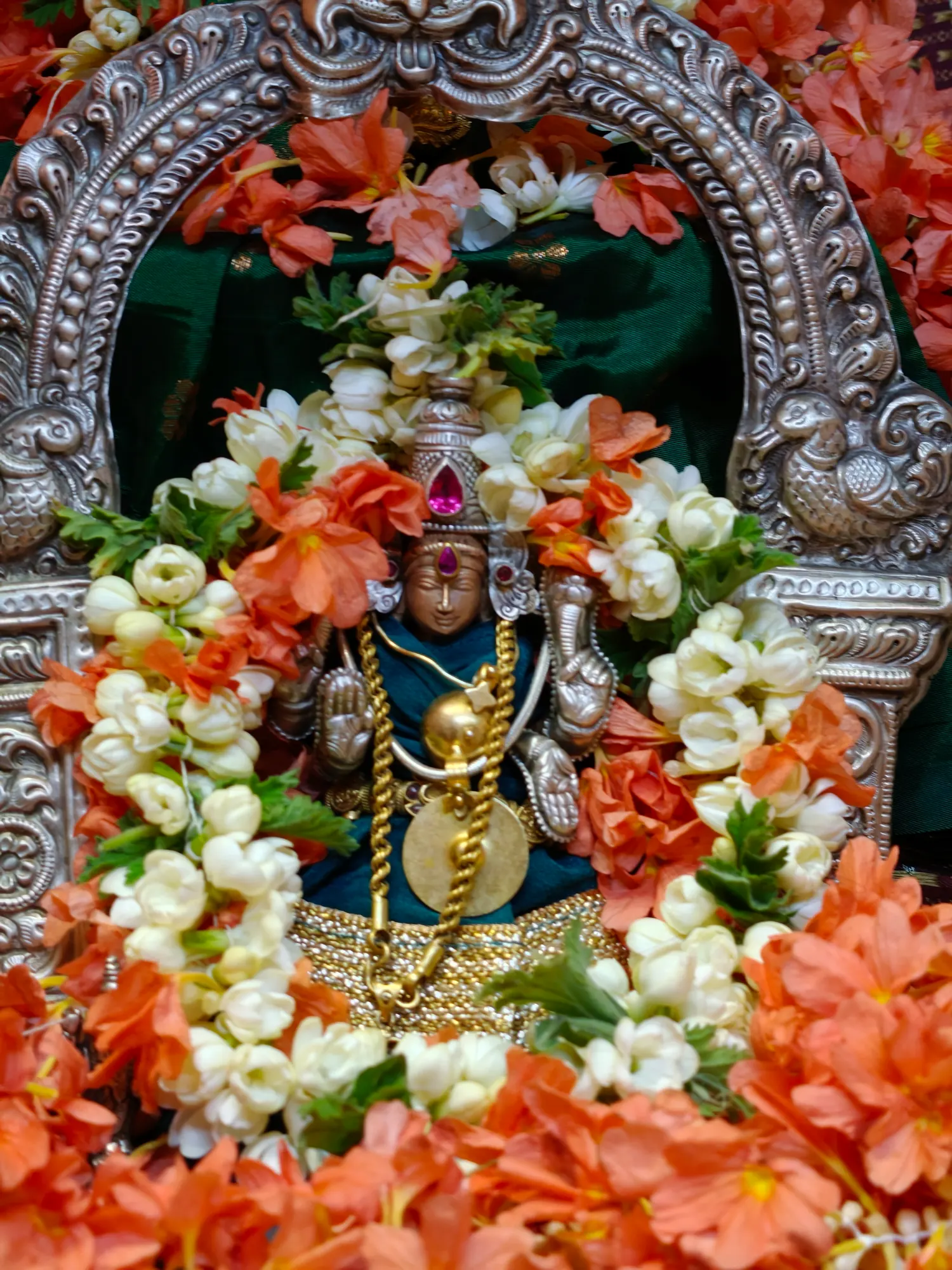 A murti seated within a silver prabhavali worked with peacocks, nearly covered over by jasmine buds and coral-coloured blossom