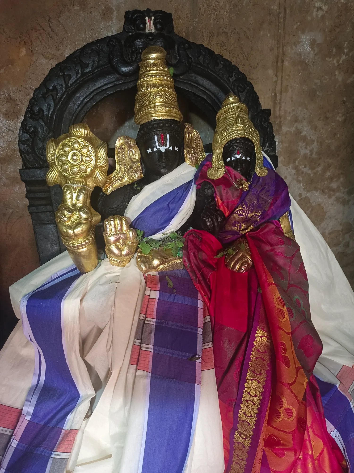 A black stone murti of the Lord with his consort set in a carved niche of the pooja mandira, crowned and armed in gold and dressed in blue-bordered white and deep red silk