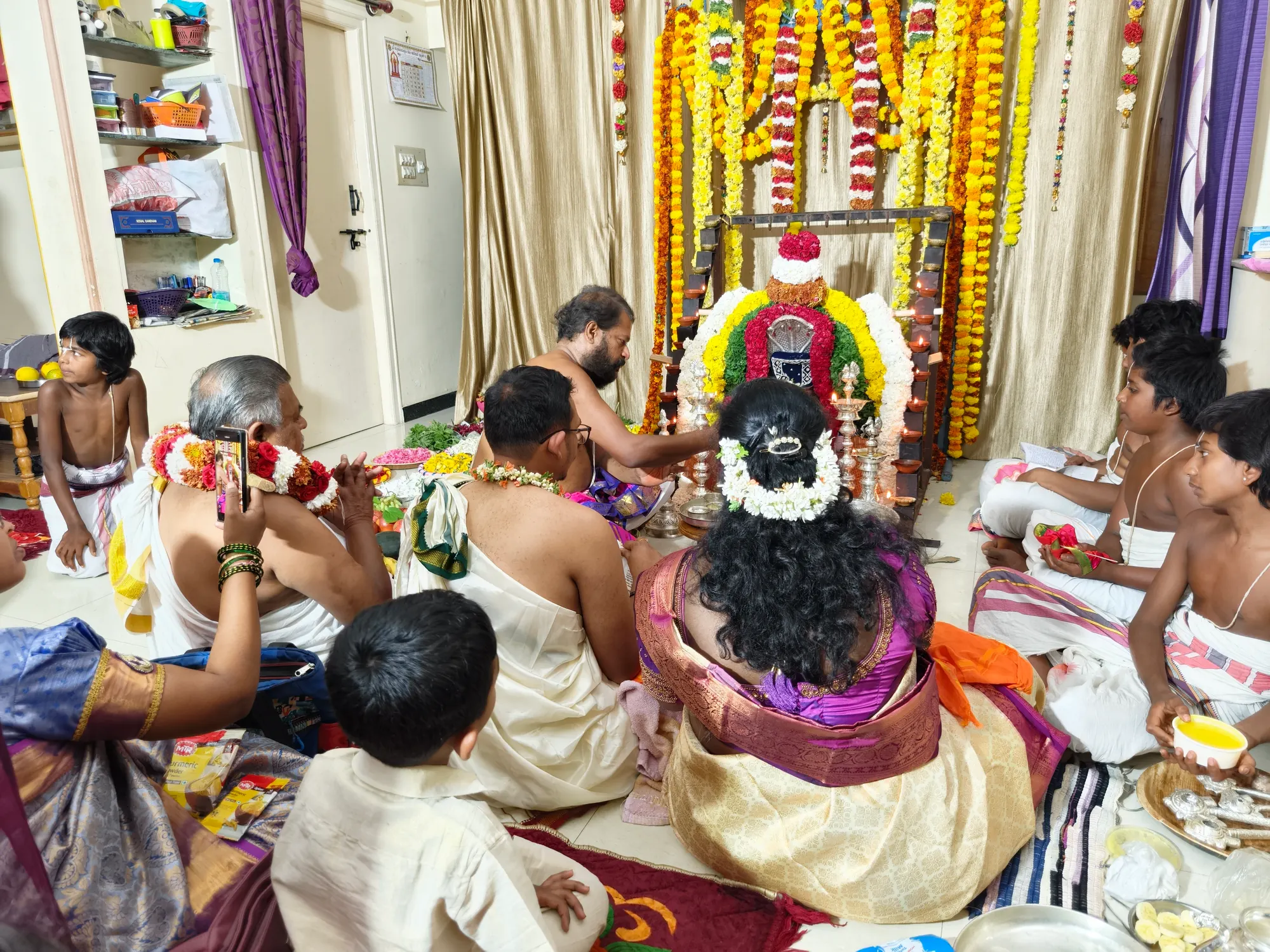 A household pooja seen from behind the family, the shrine hung with long marigold strings and the priests and children seated close around it