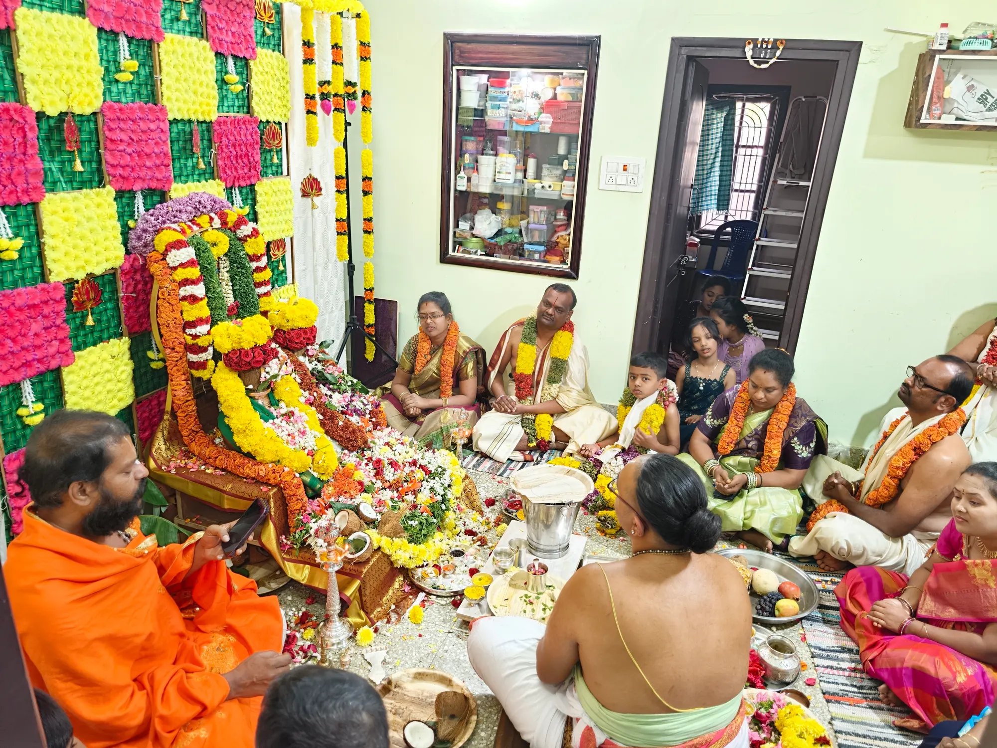 A pooja conducted in a family's own house — the deity garlanded against a flower backdrop, the family seated around the offerings and Guruji at the left
