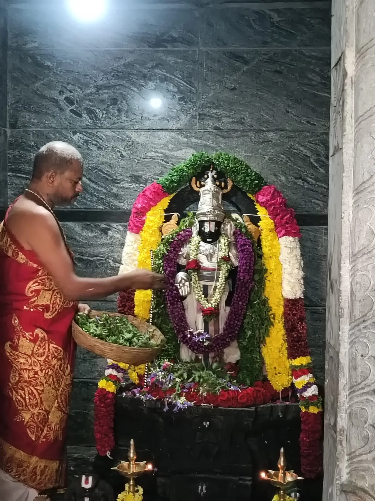 An archaka in a red-bordered silk dhoti laying tulasi from a cane basket over a newly consecrated stone murti, garlanded in white, yellow and magenta flowers against a polished granite wall, with oil lamps lit at its feet