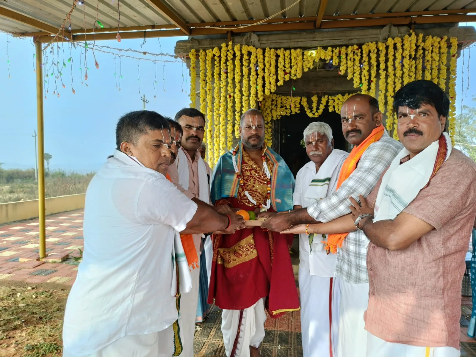 The trustees of a new temple reaching in together to hold a single offering plate with Guruji, the marigold-hung shrine open behind them under a bamboo canopy