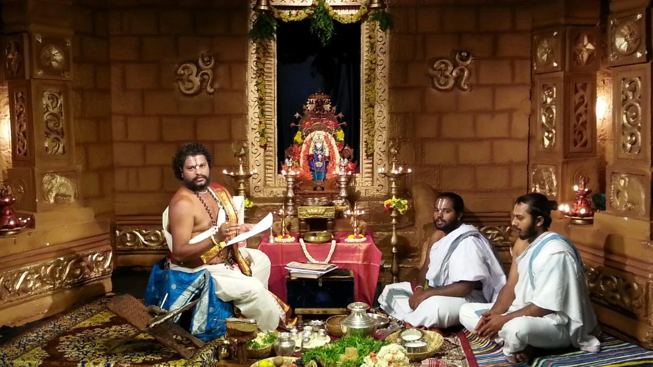 A vaidika seated cross-legged reading aloud from a bound text before a newly dressed sanctum, two students seated opposite him, the deity garlanded in its lit niche and the offering vessels set out across the floor between them