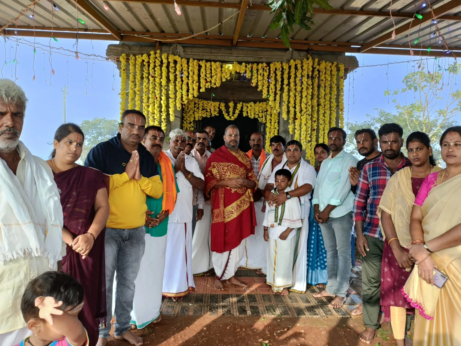 The village standing before a small stone shrine hung head to foot in marigold garlands on the morning of its consecration, Guruji at the centre in a red shawl with a child beside him