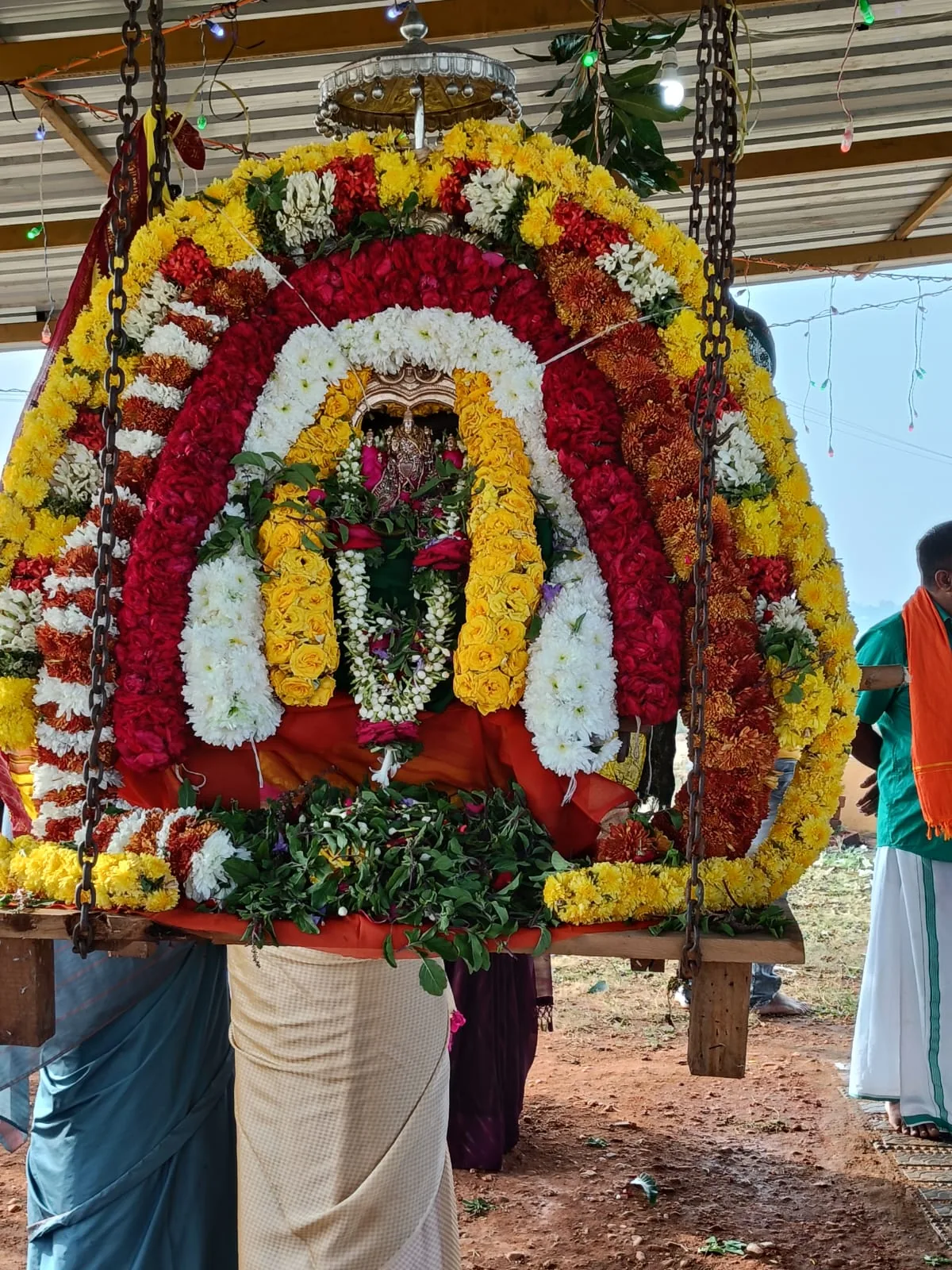 The utsava murti carried out on a wooden palanquin inside an arch of red, white and yellow flowers built around it, borne on the shoulders of men in white dhotis