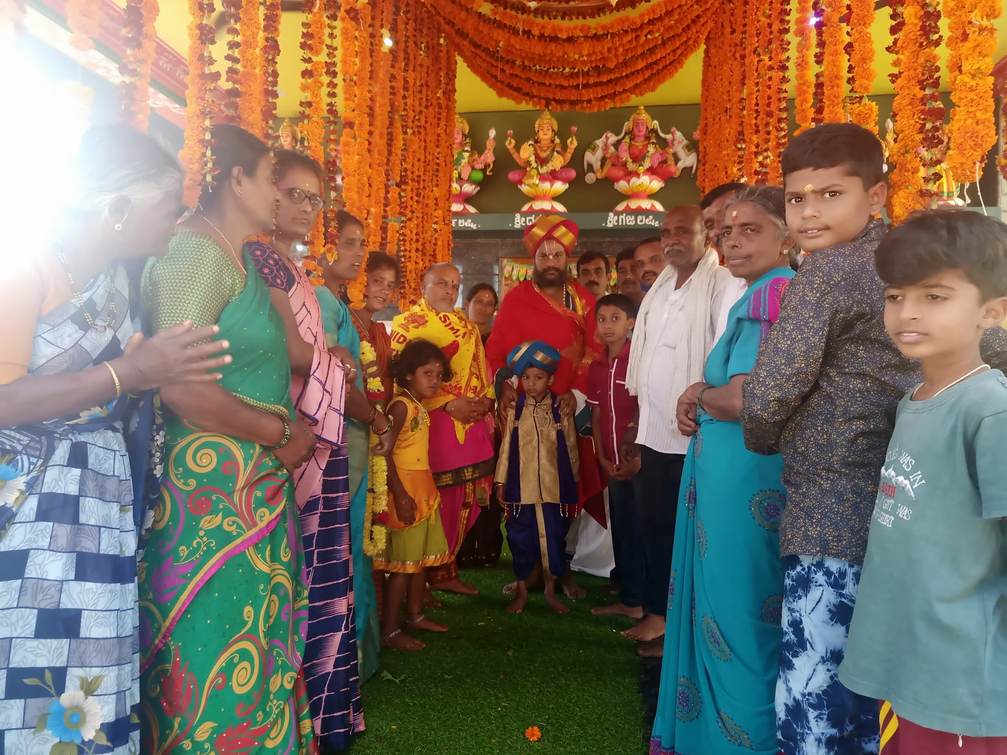 Families packed in on both sides of a shrine dressed in orange and marigold for its consecration, children carried on their parents' arms, the deities visible in the lit niche at the far end