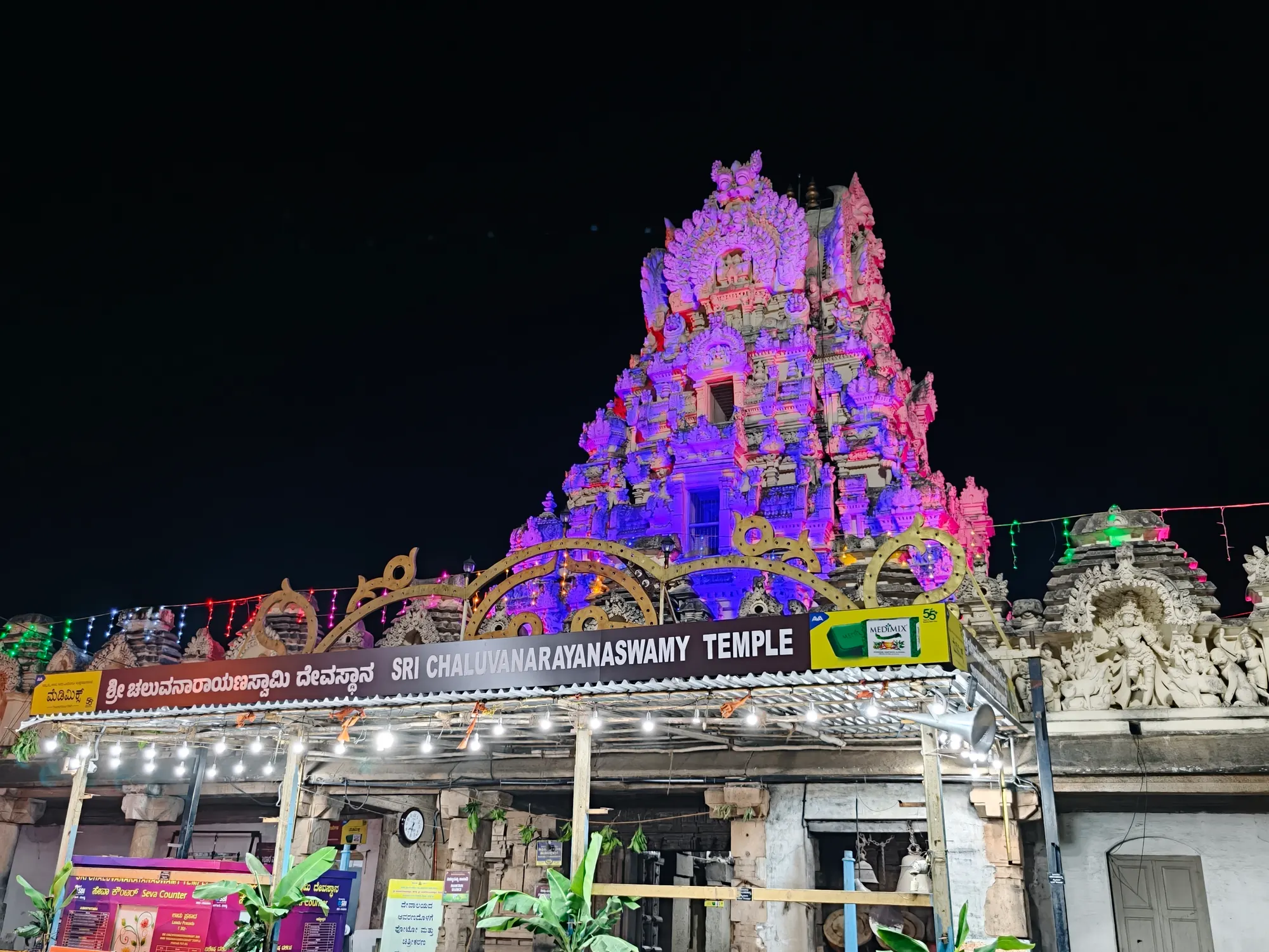 The Rajagopuram of Sri Cheluvanarayana Swamy Temple floodlit in violet and pink after dark, with the temple's name board and strings of lights along the entrance beneath it
