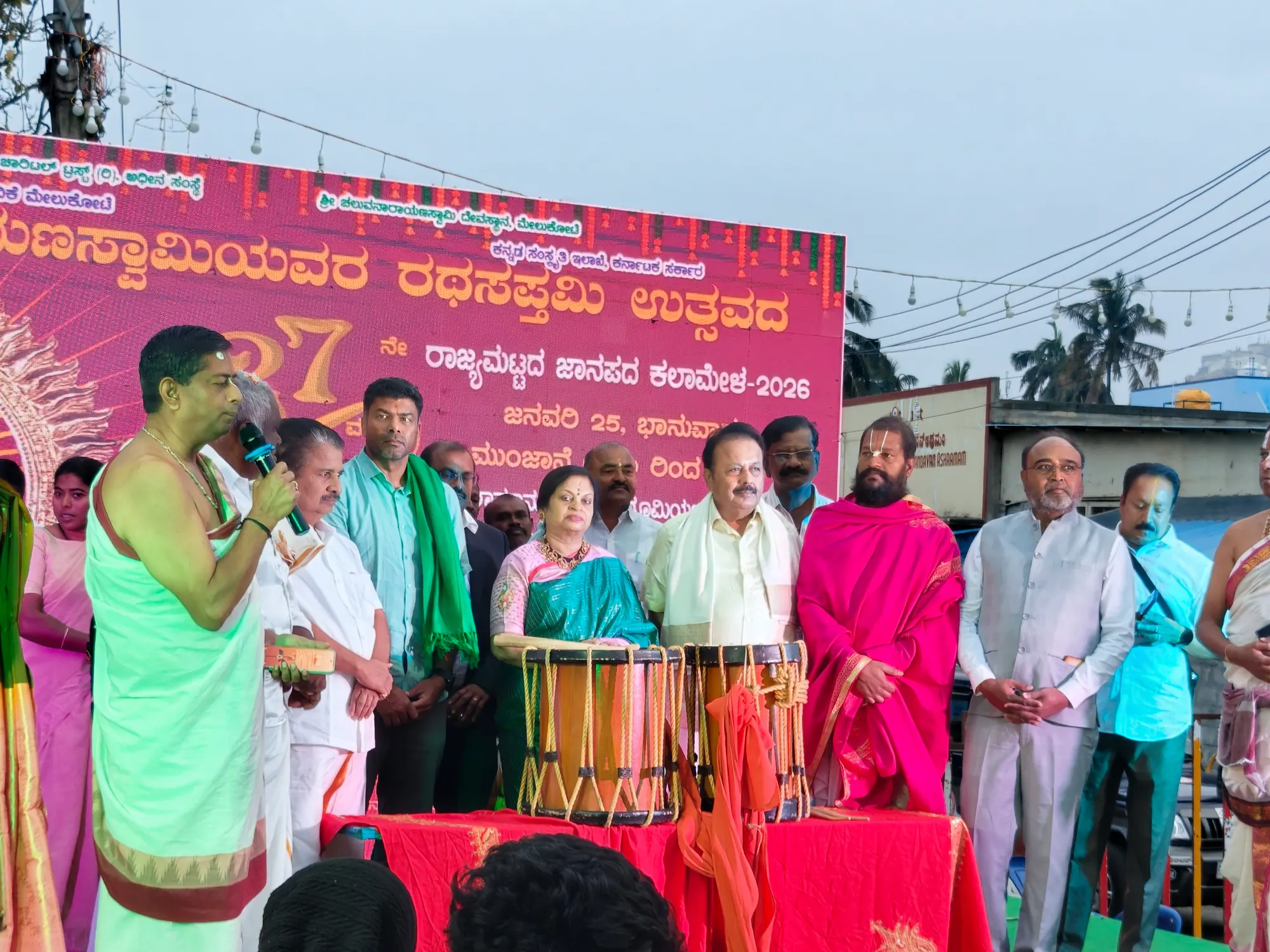 The Rathasaptami utsava stage beneath its banner for the state folk-arts mela, a priest speaking at the microphone with the drums laid out on the table before the guests