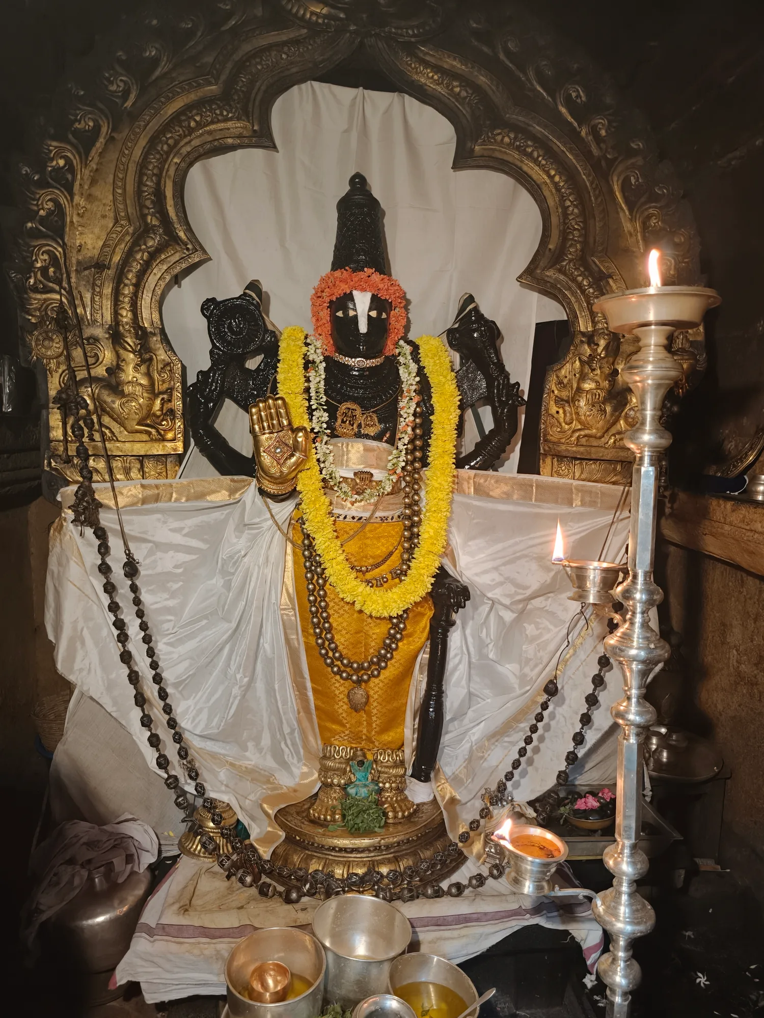 A black stone murti of the Lord in a gilded niche, garlanded in yellow and dressed in ochre silk, a tall silver lamp burning beside it