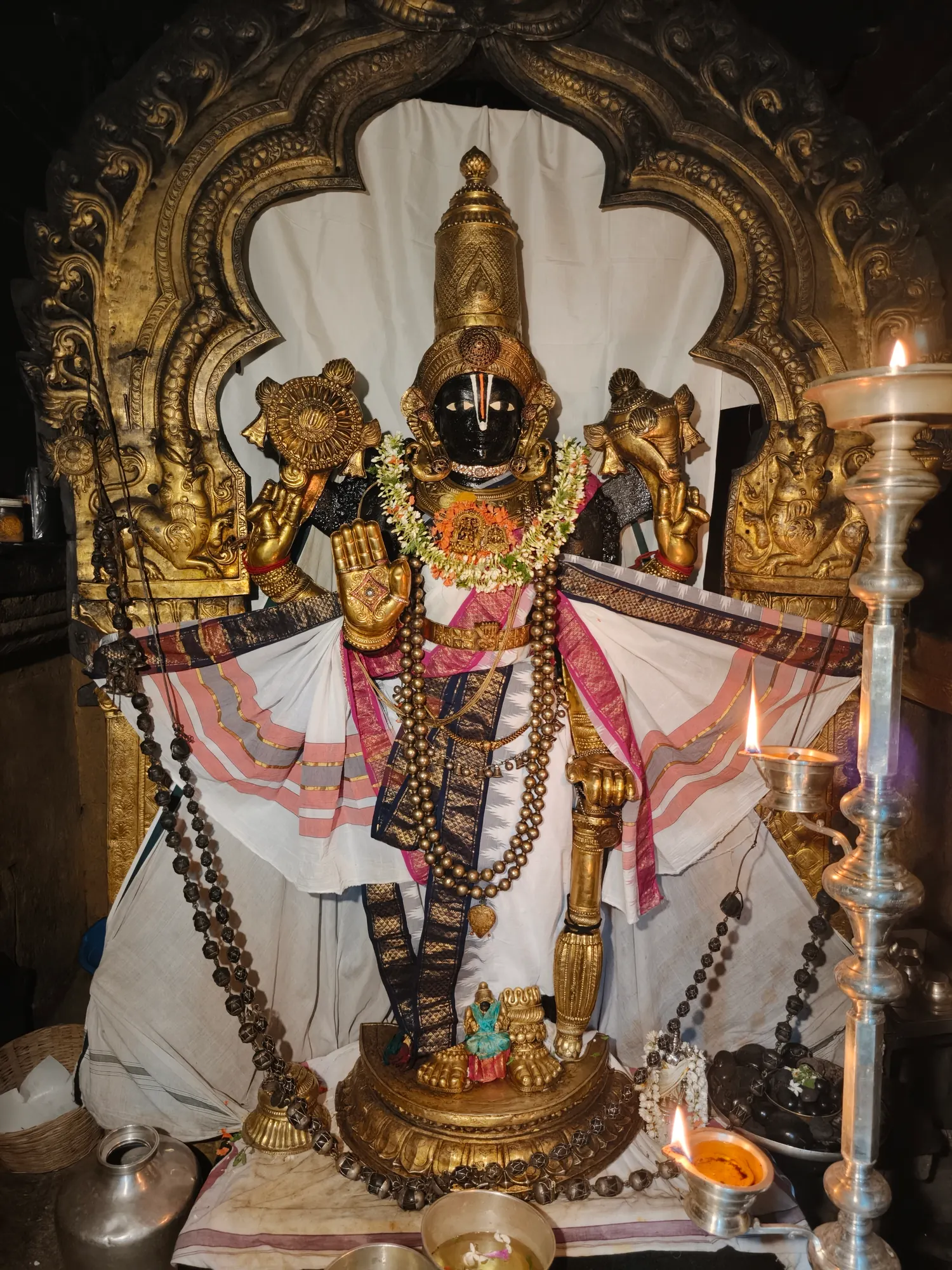 A tall bronze utsava murti of the Lord standing in a carved prabhavali, draped in white and gold cloth and hung with long sandalwood bead garlands, lit by oil lamps at either side