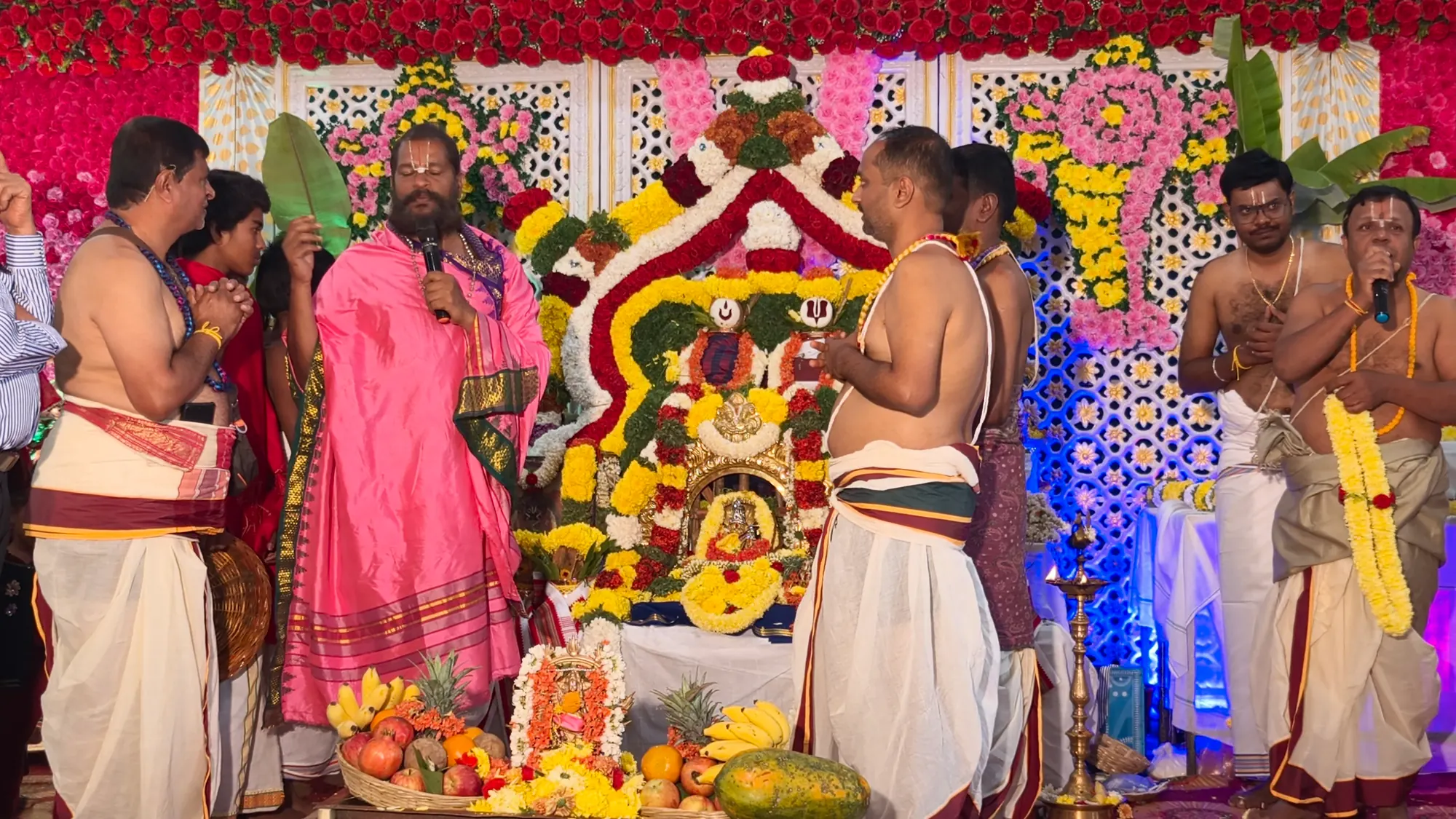 The utsava murti banked in flowers on the festival stage, Guruji speaking at the microphone with the temple's priests standing around it