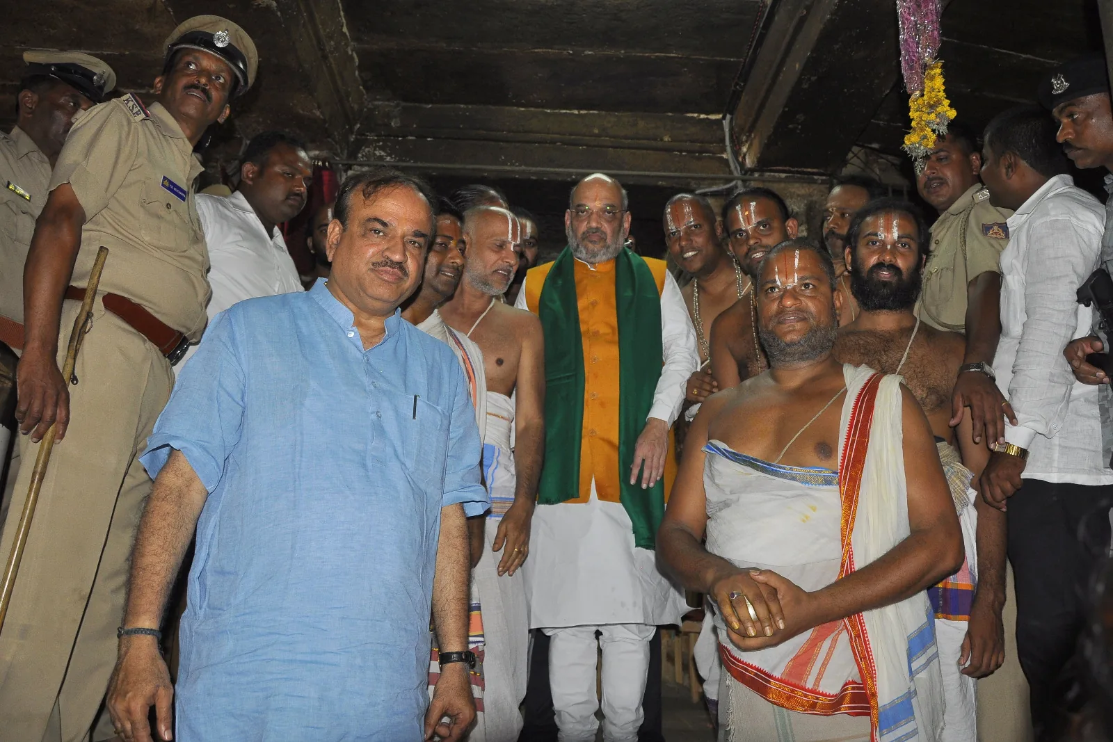 Amit Shah and Ananth Kumar standing with the archakas of the temple in the dim of the inner corridor after darshana, the police escort ranged along the wall behind them