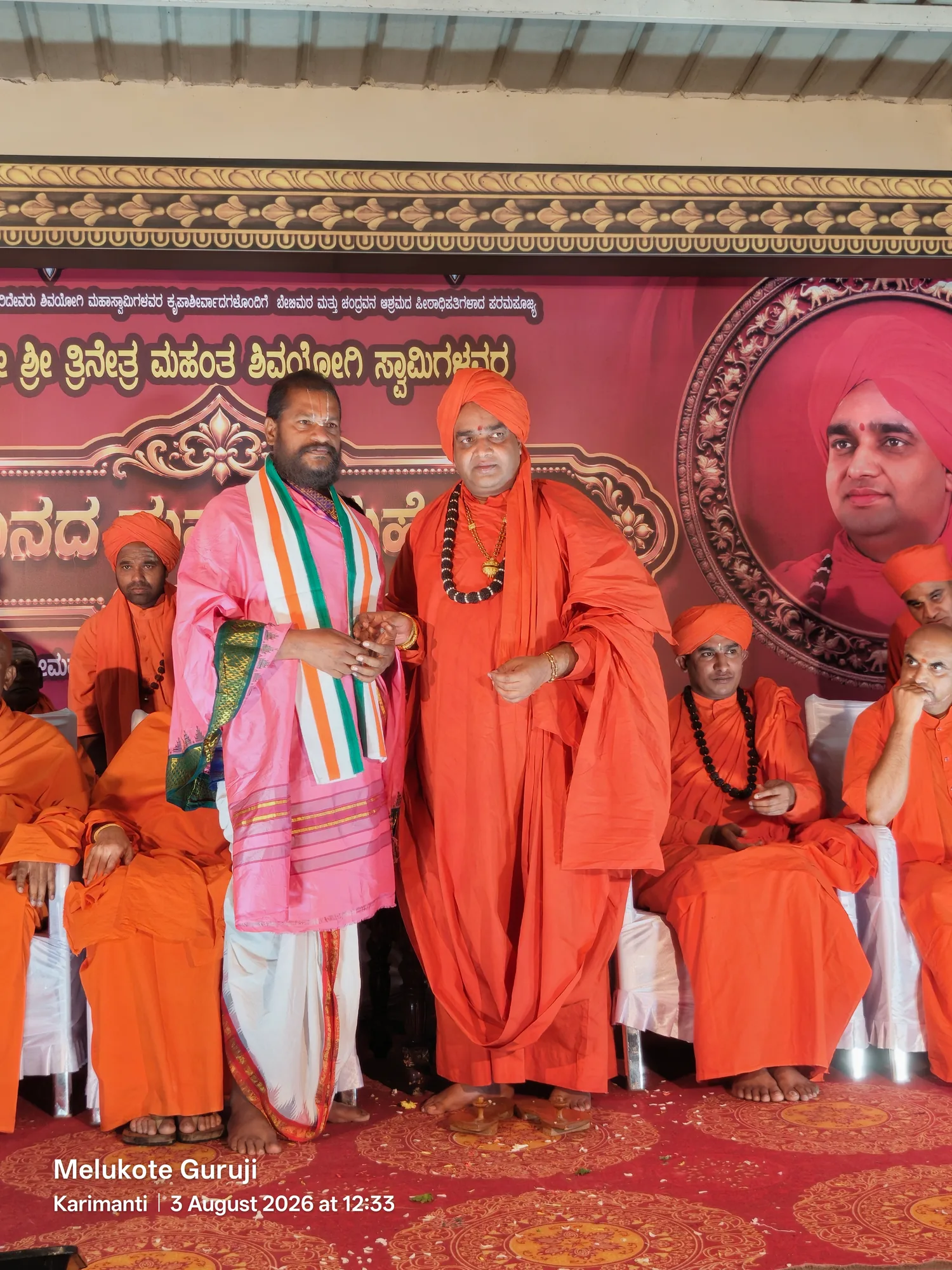 Guruji in a rose shawl shaking hands with the Swamiji on a function stage, the mutt's saffron-robed sanyasis seated on either side of them beneath the banner