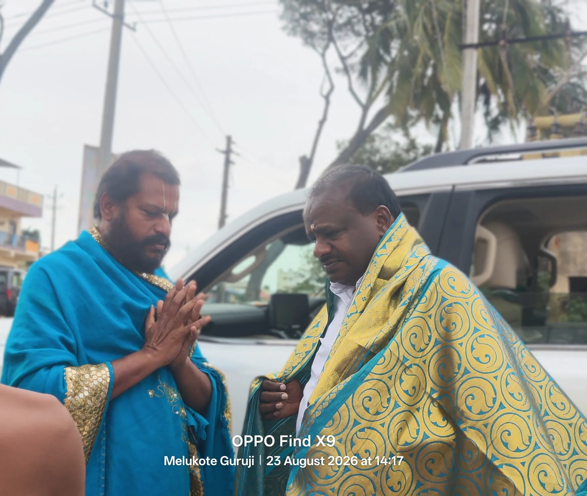 H. D. Kumaraswamy wrapped in a yellow and green shawl, head bowed, being received in the street by Guruji in blue silk with his palms joined