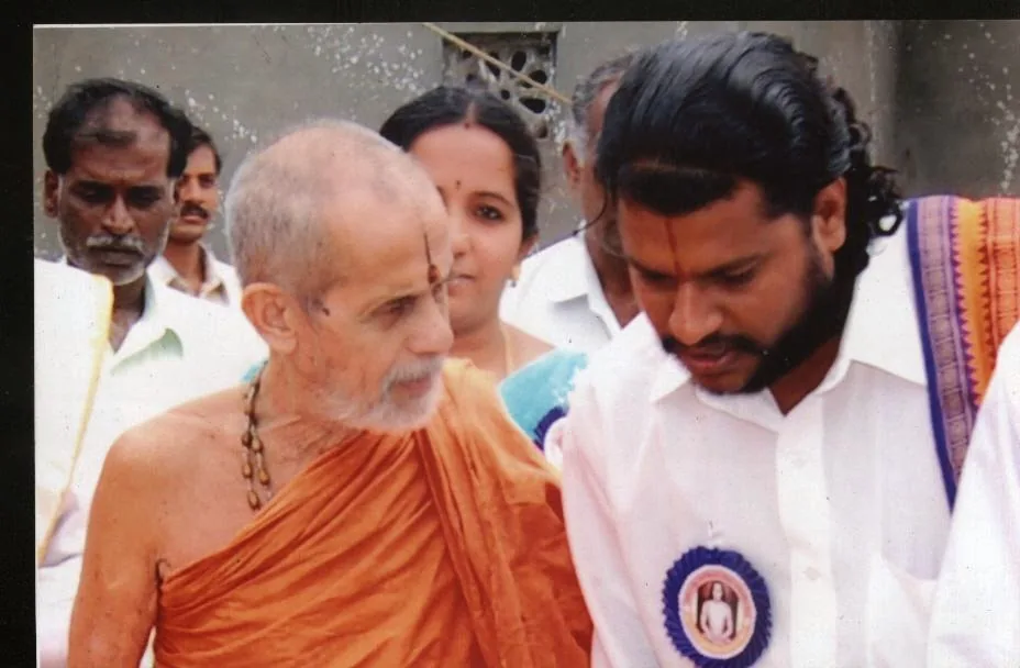 The Pejavara Swamiji in ochre robes walking out through a crowd at the kshetra, a young Guruji at his side wearing the badge of the day's function