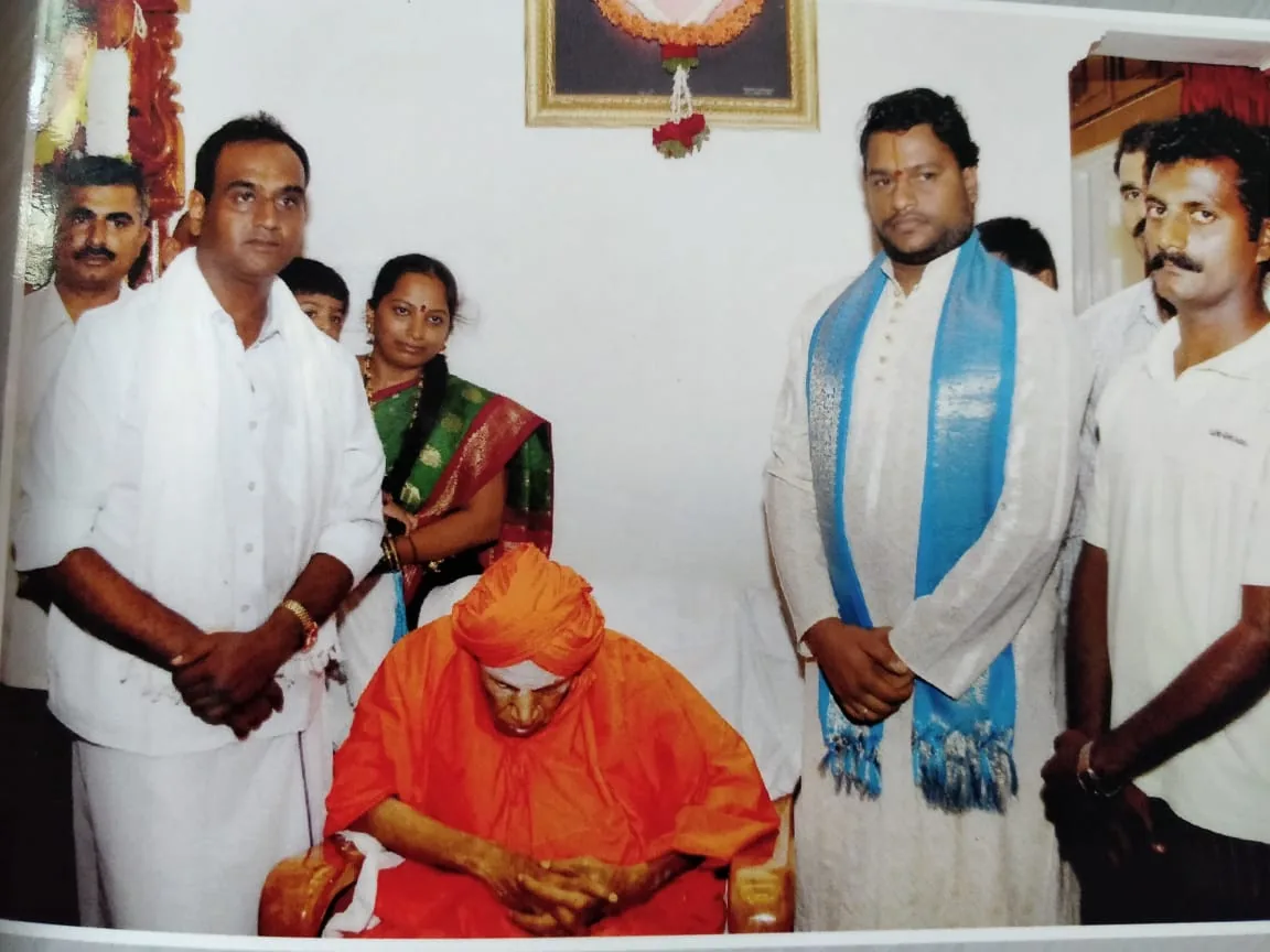 Sri Siddaganga Swamiji seated in saffron with his head bowed, the hosts of the function standing on either side of him — a print photograph from the trust's own album