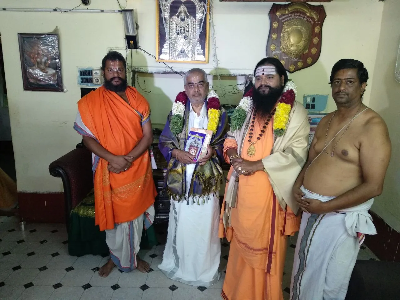 The head priest of Tirupati, garlanded and holding a book presented to him, standing between Guruji in saffron and two vaidikas of the mandira