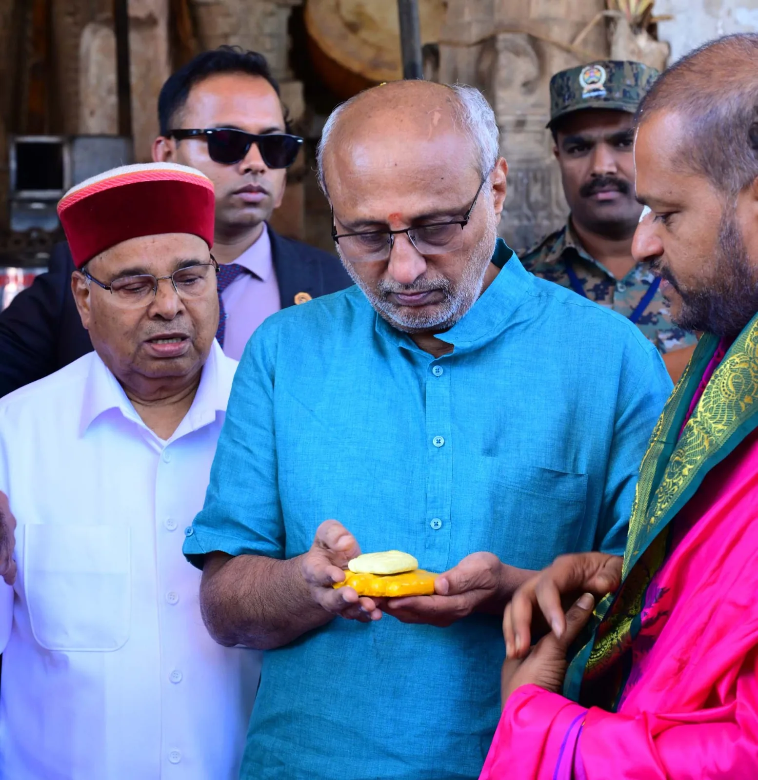 The Vice-President of India taking prasada in his cupped hands from the archaka at the temple, an aide in a maroon cap at his shoulder and his security detail behind him