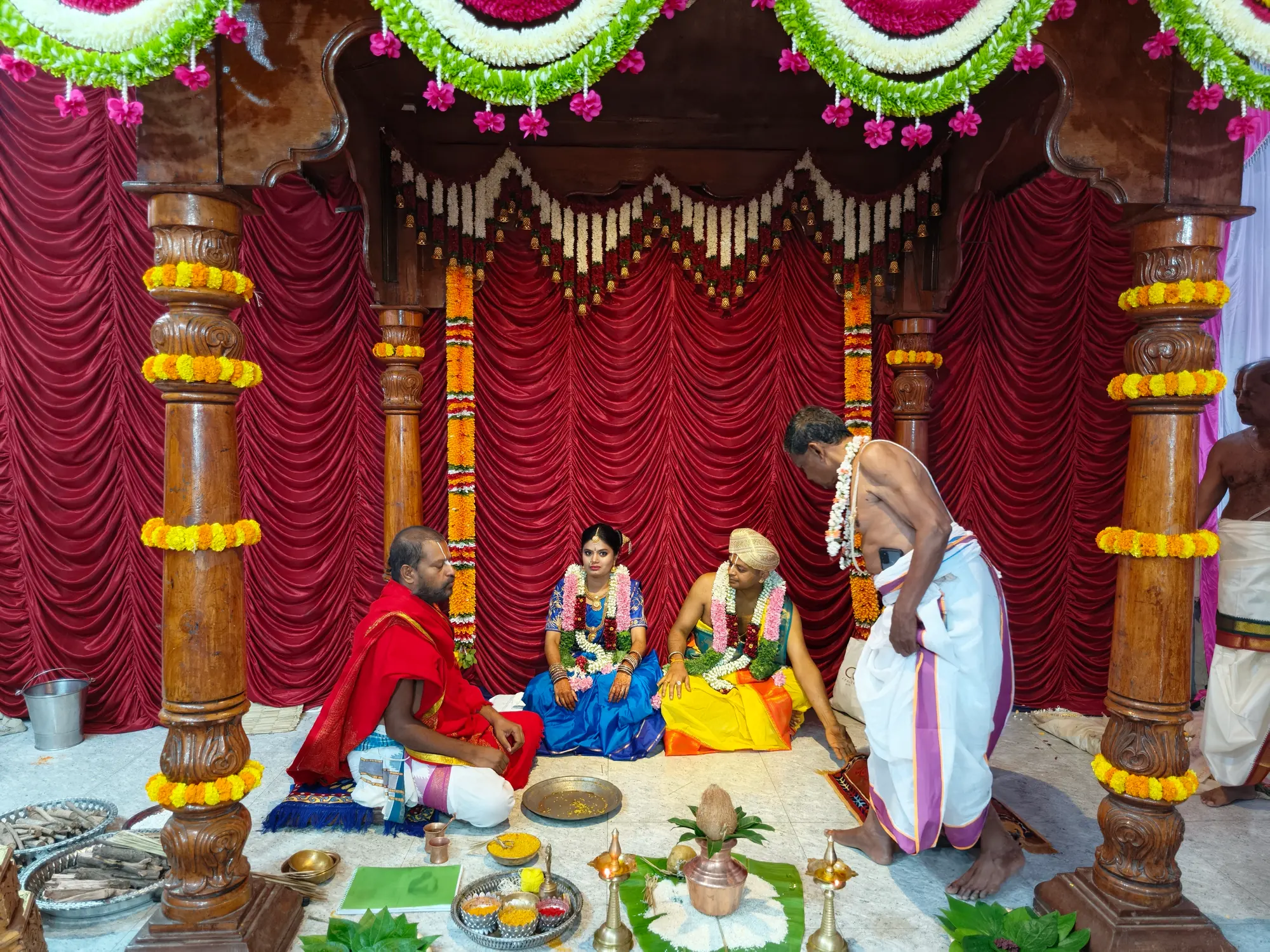 The bride and groom seated in the mandapa against a red curtain while the purohita in red conducts from the left and an assistant stands at the right