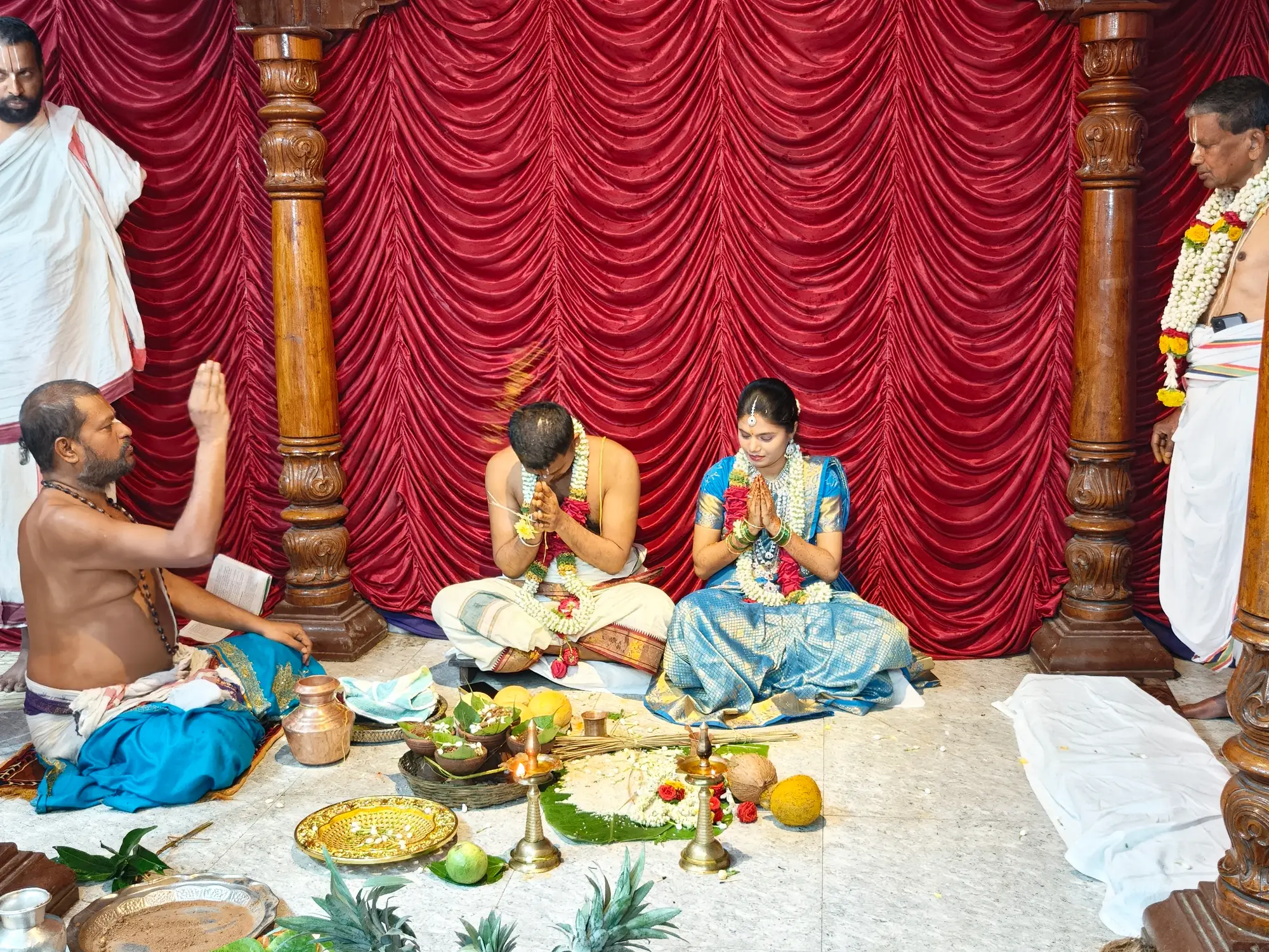 The bride and groom seated in the mantapa with palms joined, the purohita conducting from the left and the offerings set out between them