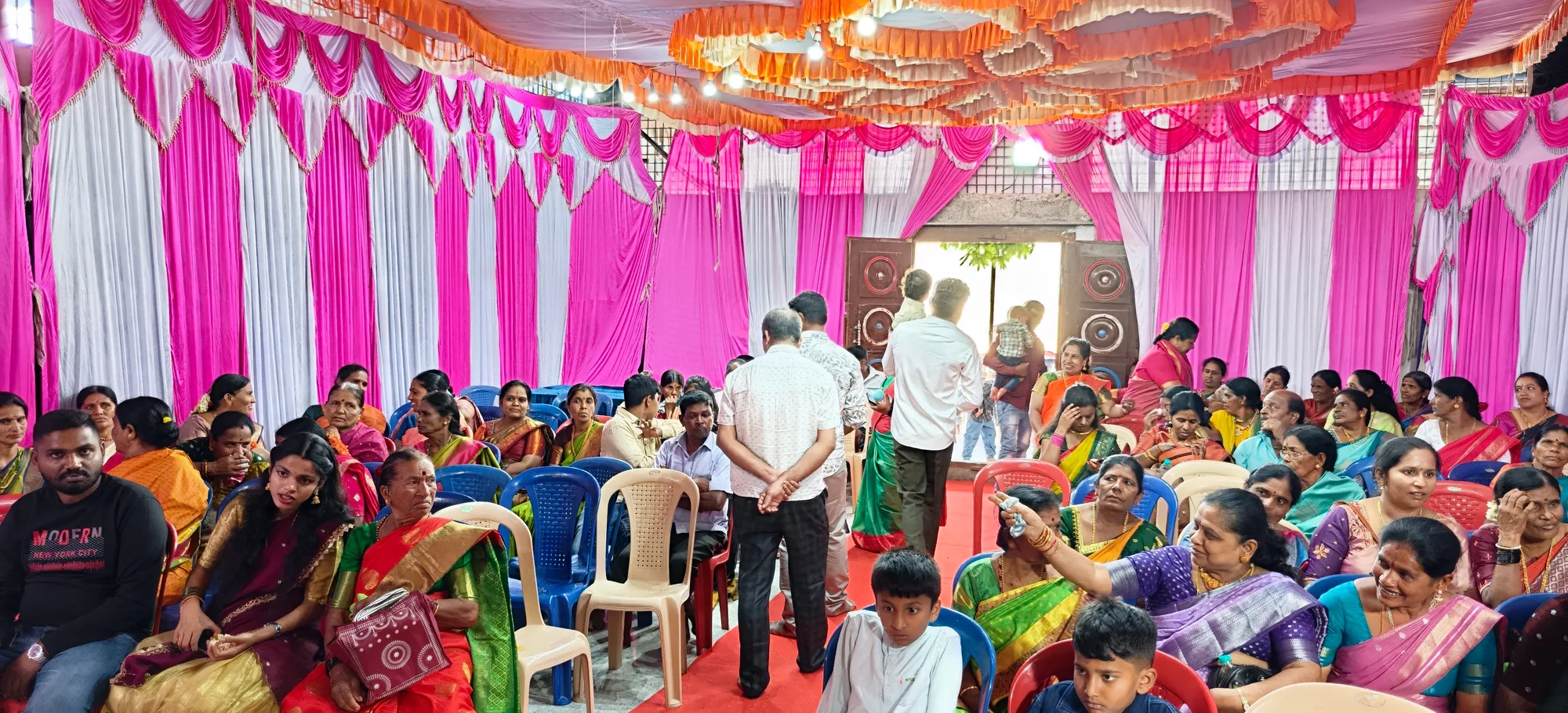 The whole hall of guests seen from the back, children on the floor at the front and the decorated stage beyond them