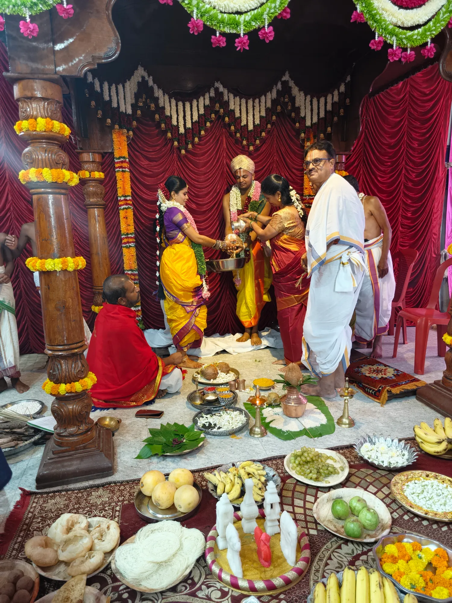 The groom standing in the mandapa while the women of the family hold the kalasha between them, the purohita seated at the left and the offering plates set out in front