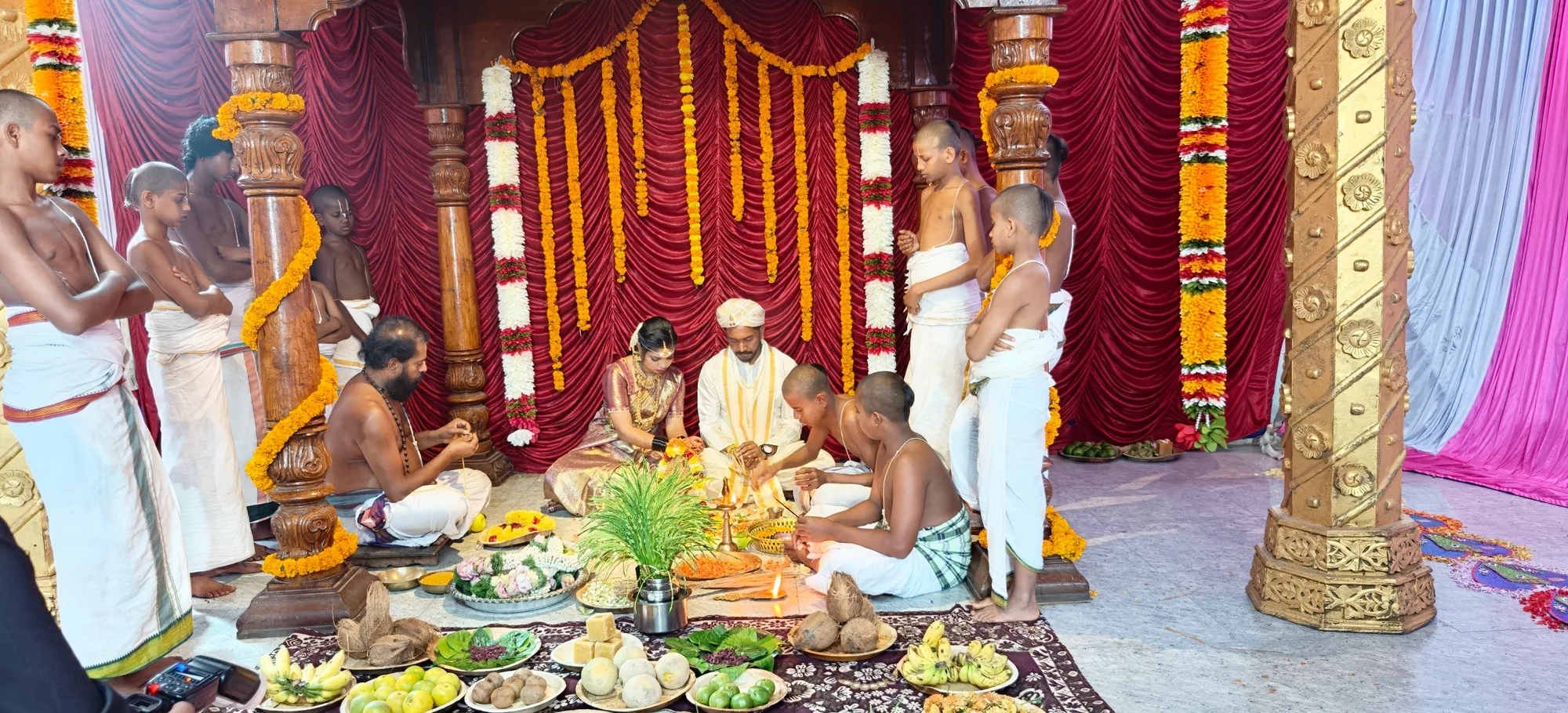 The mandapa seen across the width of the hall, the couple seated for the sacred rituals of the rite, and the offerings laid the whole way along the front
