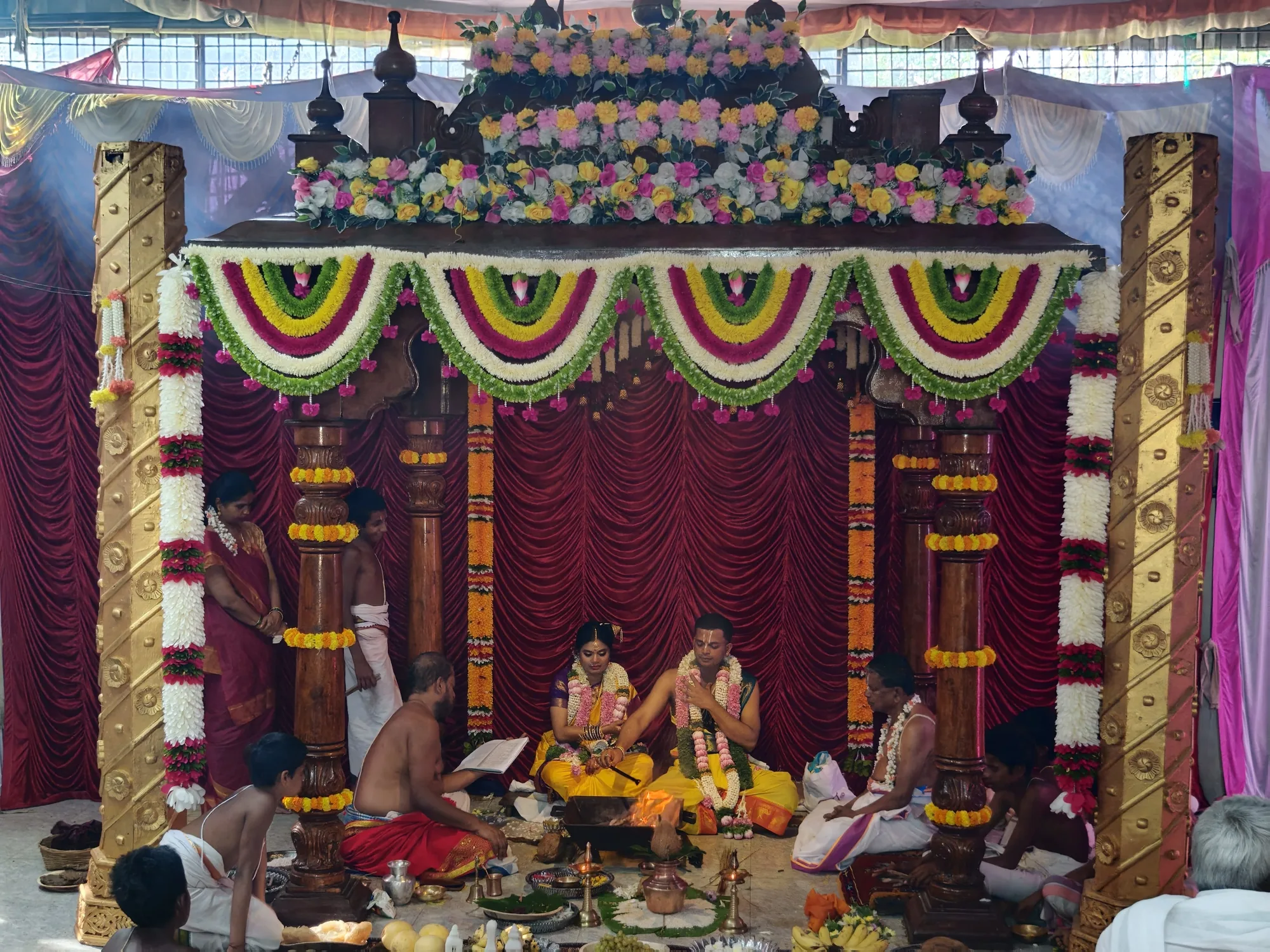 The mandapa seen from the hall floor — the couple seated at the centre with the purohitas around them, the whole frame hung with flowers