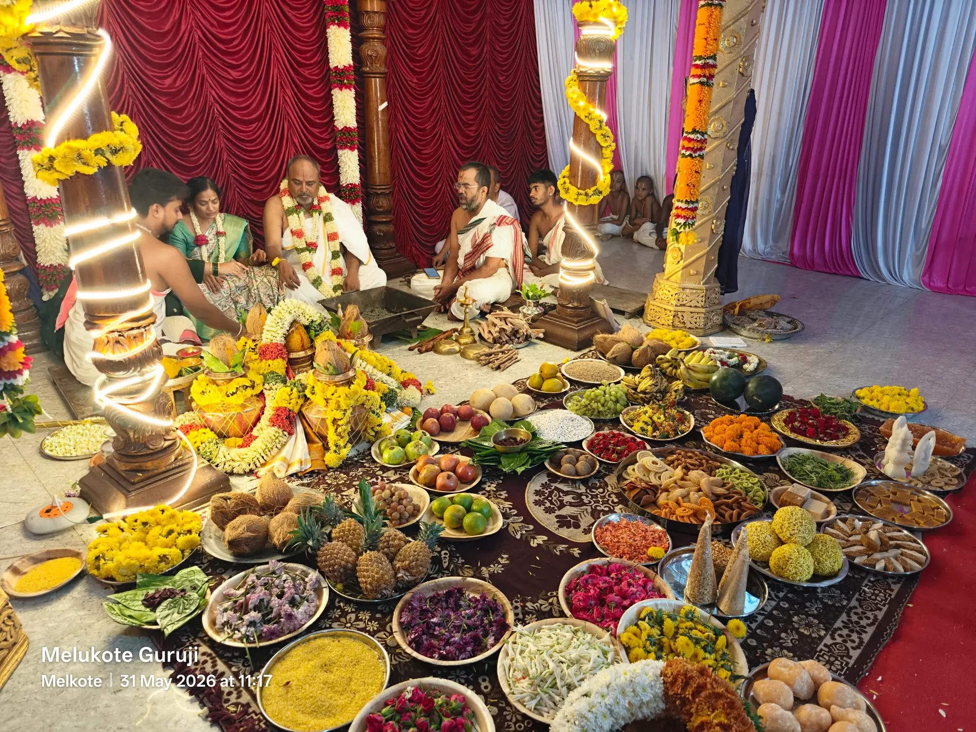 The wedding offerings seen from above — pineapples, oranges, sweets and heaped flowers in metal dishes covering the carpet in front of the mandapa