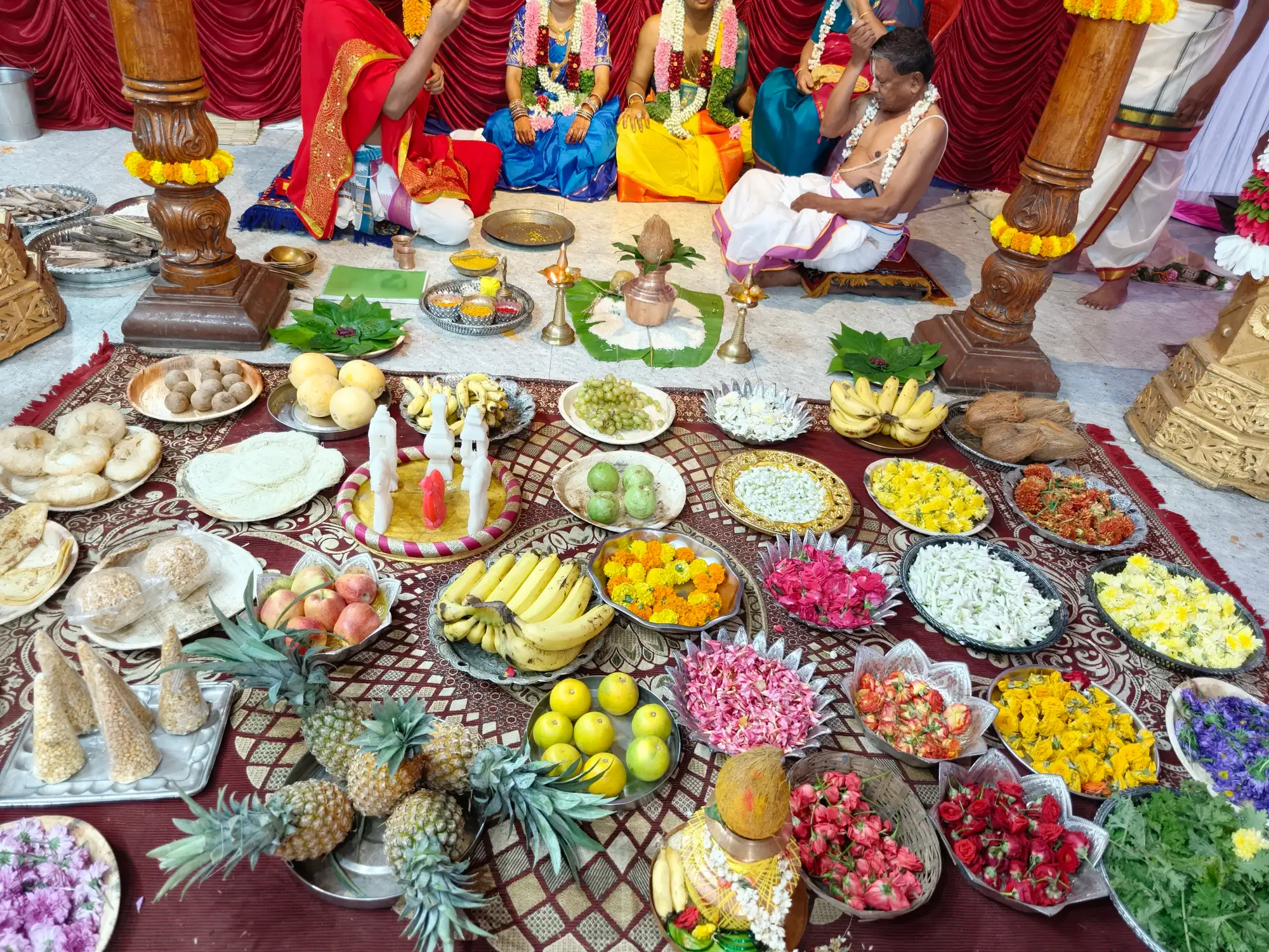The wedding offerings laid out on a cloth across the floor — pineapples, bananas, apples and limes in metal dishes among trays of loose flowers and sweets