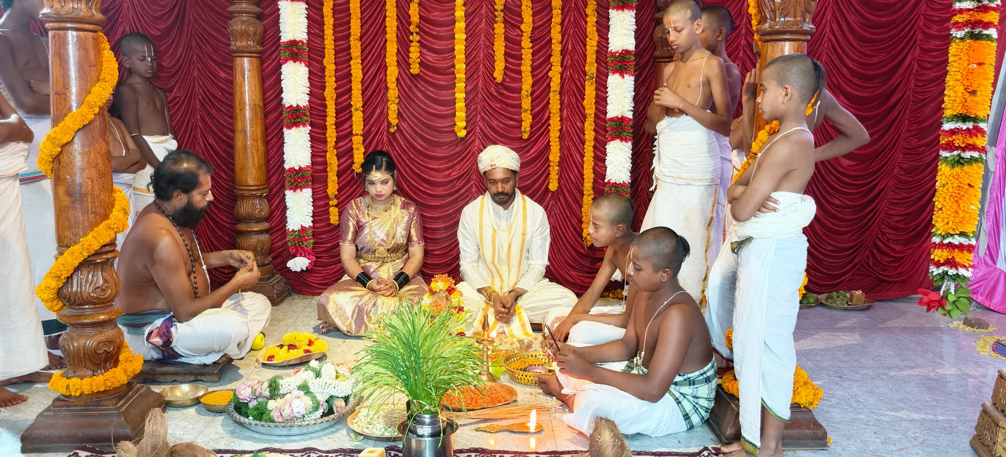 The couple seated in the mandapa with the purohita and the young students assisting him, in front of red drapes and marigold-wound pillars