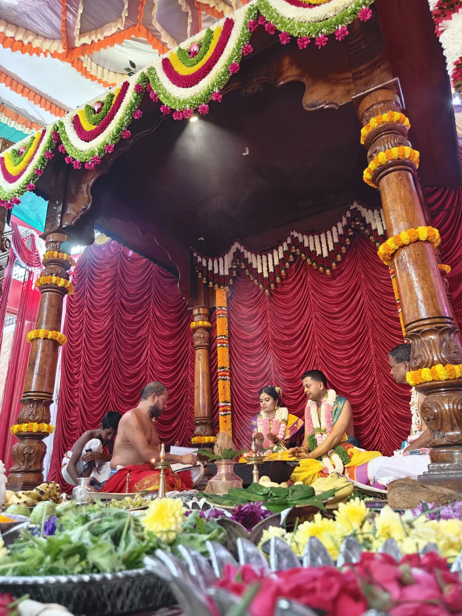 The couple seated in the mandapa under a canopy of pink and green garlands, the purohita conducting at their side and the offerings filling the foreground