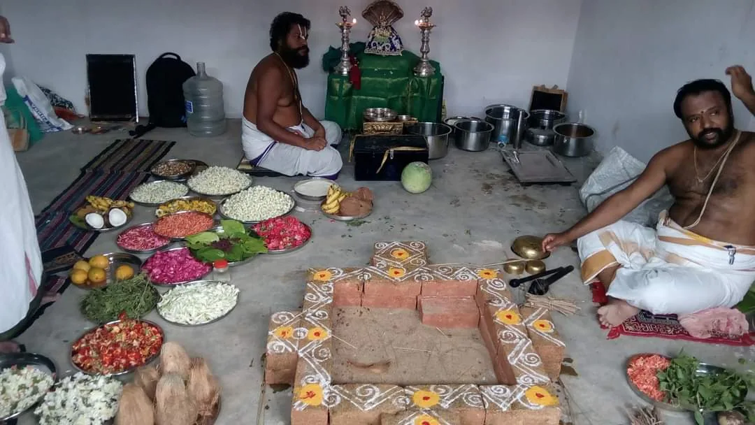 Two priests seated on either side of a freshly built brick homa kunda drawn round with rangoli, the whole spread of the day's offerings laid out in dishes across the floor between them — cut fruit, flowers, greens and grain