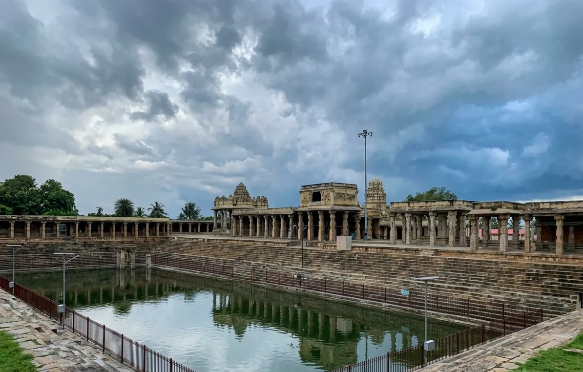 The kalyani at Melukote (Melkote) under a heavy grey monsoon sky, its green water fenced and held in broad stone steps, with pillared mandapas and two small temple towers standing along the far side.