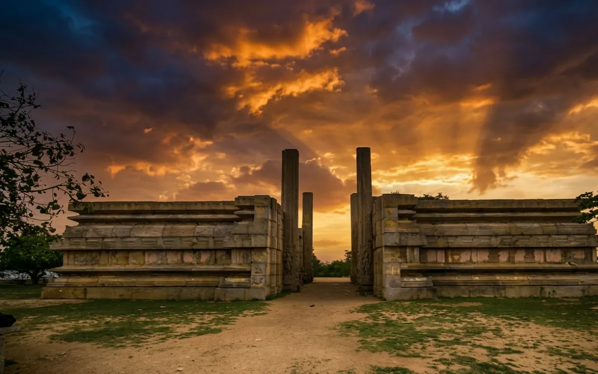 The unfinished Raya Gopura at Melukote (Melkote) against a fiery sunset: two long stone platforms in open ground with tall bare pillars rising on either side of the gap between them, and a dirt path running through.